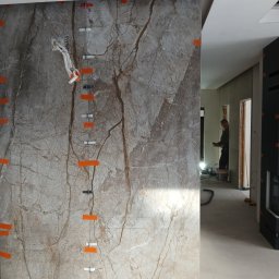 BCS - Montaż marmurowych płytek na ścianie z widocznymi elementami poziomowania i gniazdkiem elektrycznym. W tle widoczna postać pracownika.