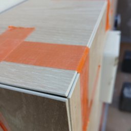 BCS - Wykończona obudowa wanny z płytek imitujących drewno, zabezpieczona pomarańczową taśmą malarską. Widoczne fugi i krawędzie płytek.