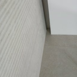 BCS - Ściana wyłożona jasnymi, prążkowanymi płytkami ceramicznymi, z widocznym fragmentem sufitu i podłogi z płytek w odcieniach beżu. Minimalistyczne wykończenie.