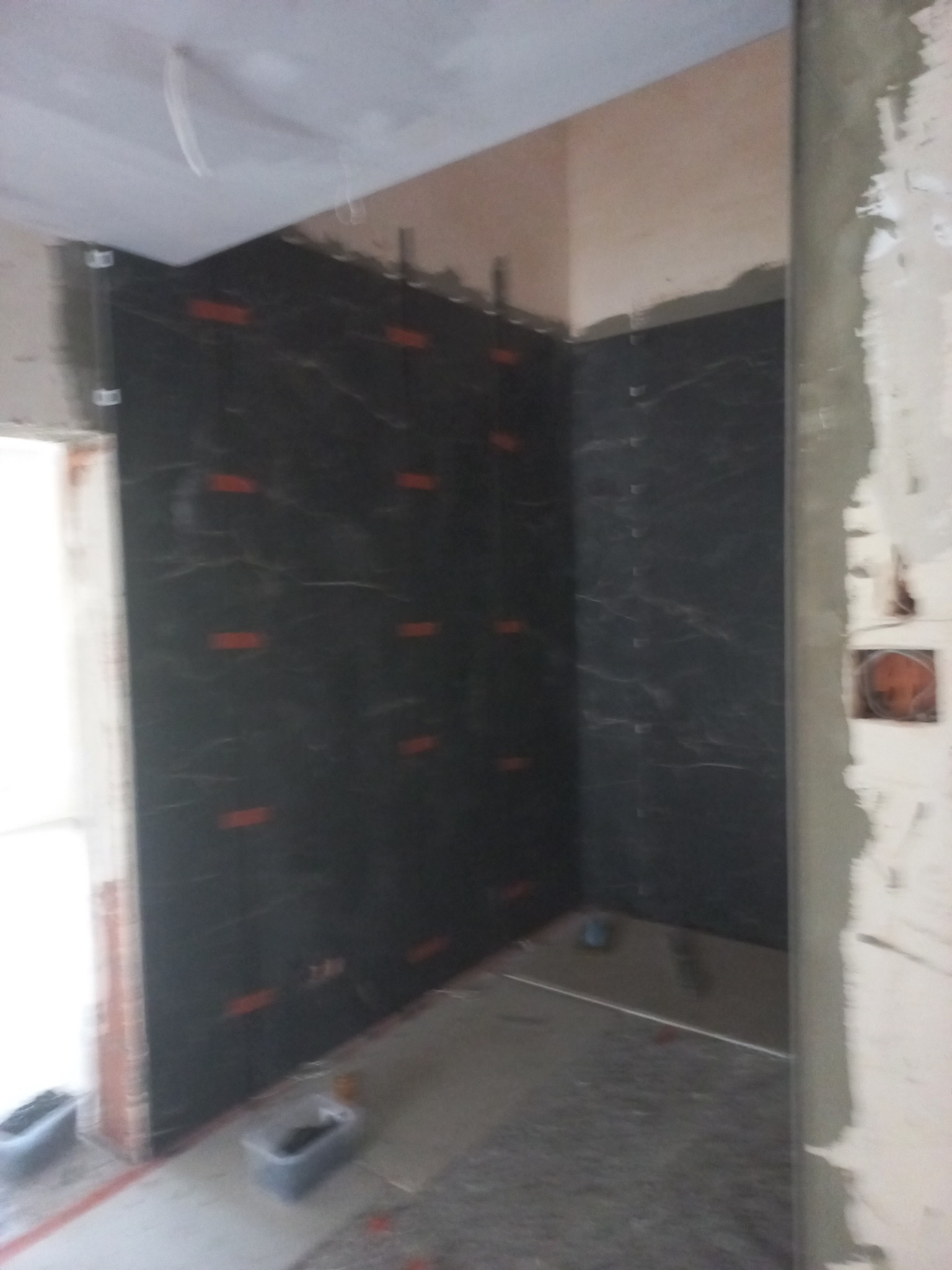 Ciemne płytki imitujące marmur, ułożone na ścianie w rogu pomieszczenia, z widocznymi elementami montażowymi i fragmentem wylewki podłogowej. Prace wykończeniowe w toku.