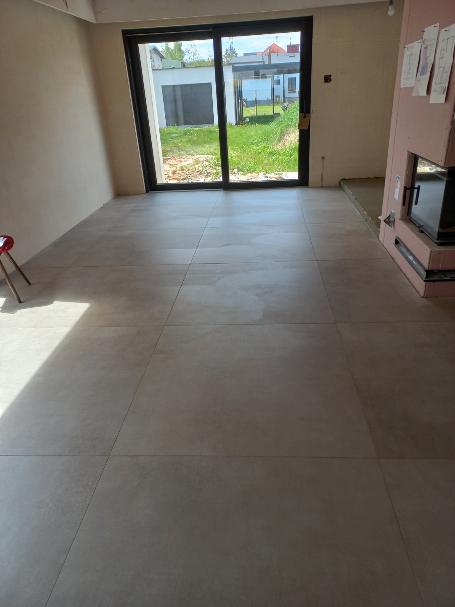 Beżowe płytki podłogowe ułożone w nowym domu, widok na przeszklone drzwi tarasowe i ogród. Fragment kominka z obudową. Minimalistyczny design wnętrza.