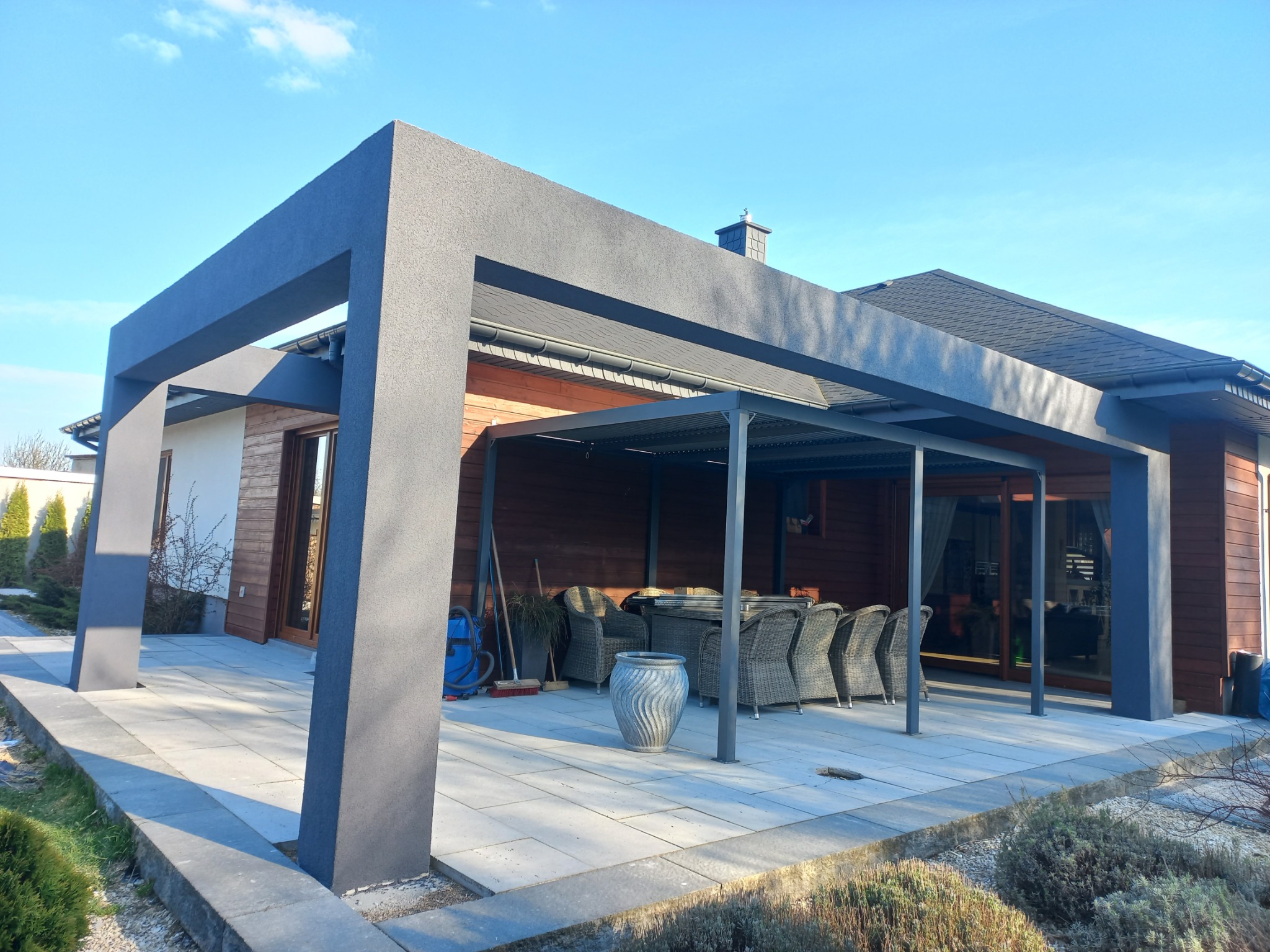 Nowoczesna pergola z szarej konstrukcji, zadaszona, z meblami ogrodowymi w tle, na tle domu z drewnianą elewacją i błękitnego nieba. Stonowana kolorystyka.