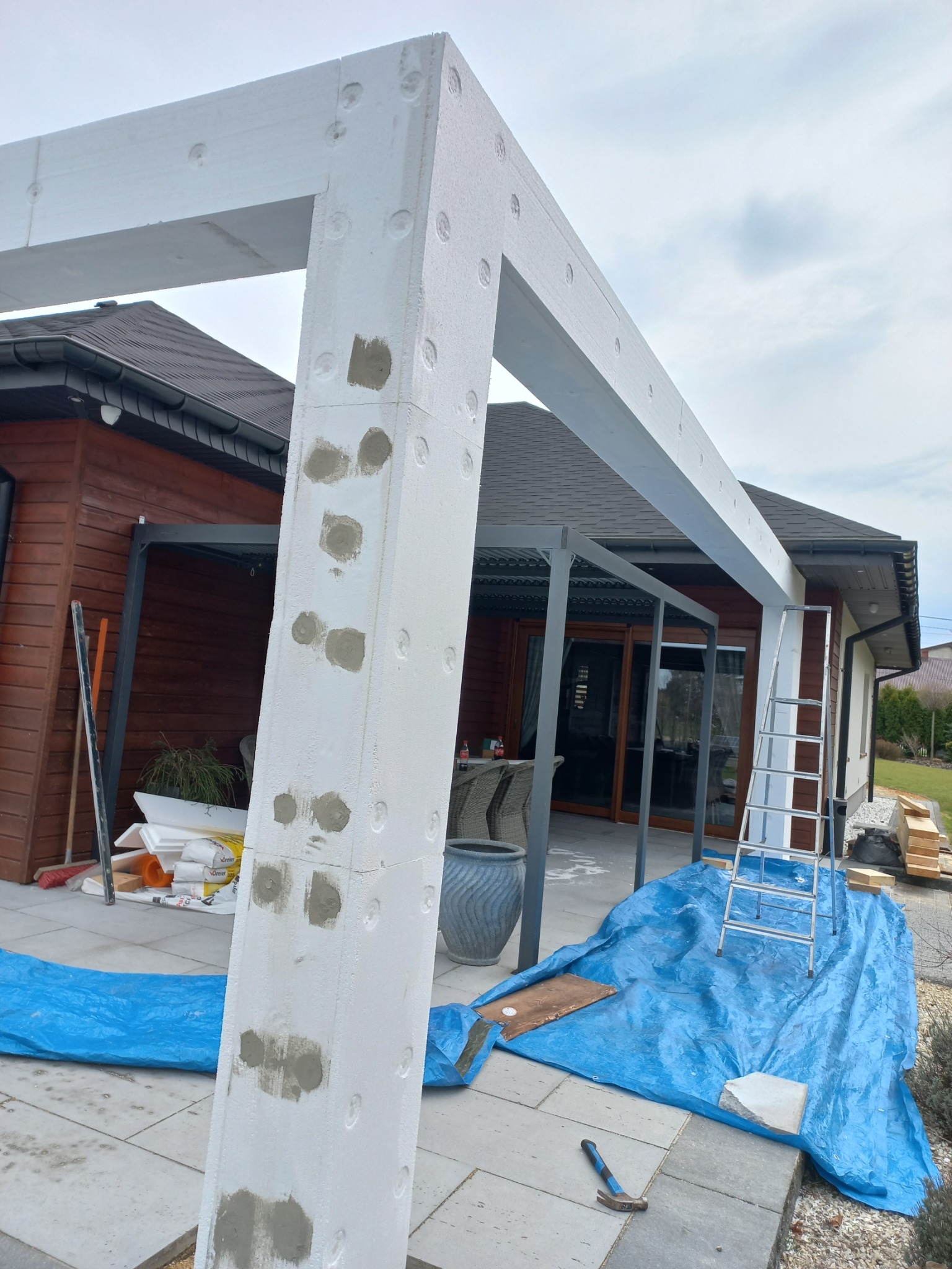 Konstrukcja pergoli z białych elementów, z widocznymi śladami szpachlowania, na tle domu z drewnianą elewacją i niebieską plandeką.