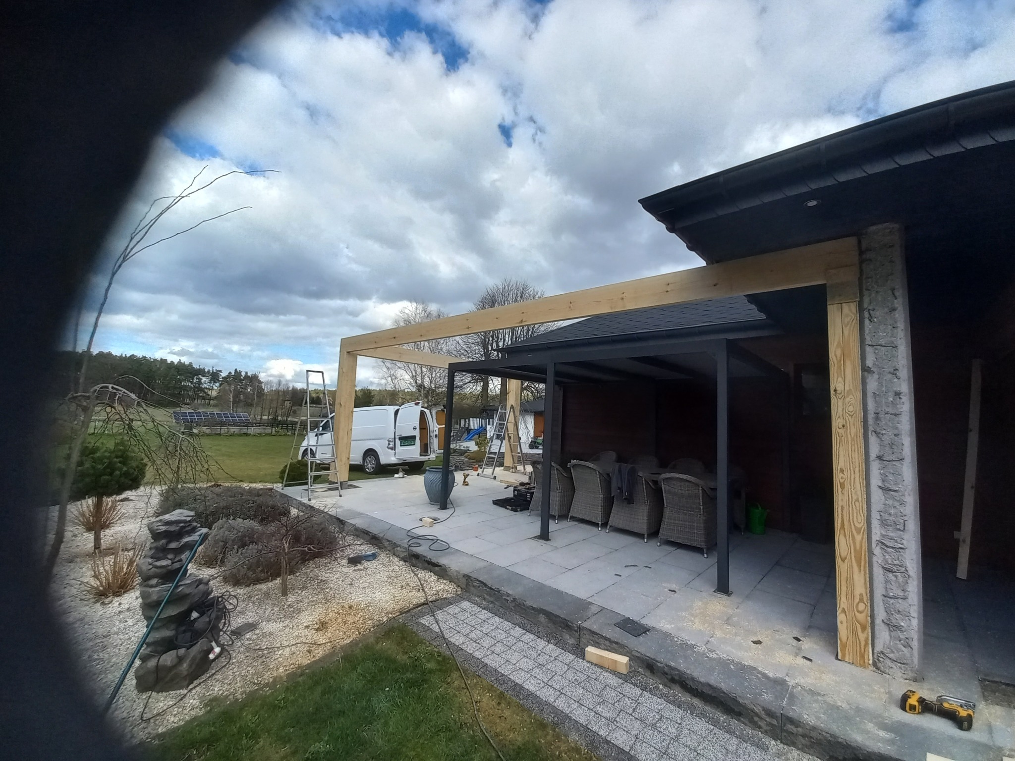 Budowa drewnianej pergoli przy domu z szarymi płytami tarasowymi. W tle biały bus i drabina. Widoczne narzędzia i elementy konstrukcyjne.