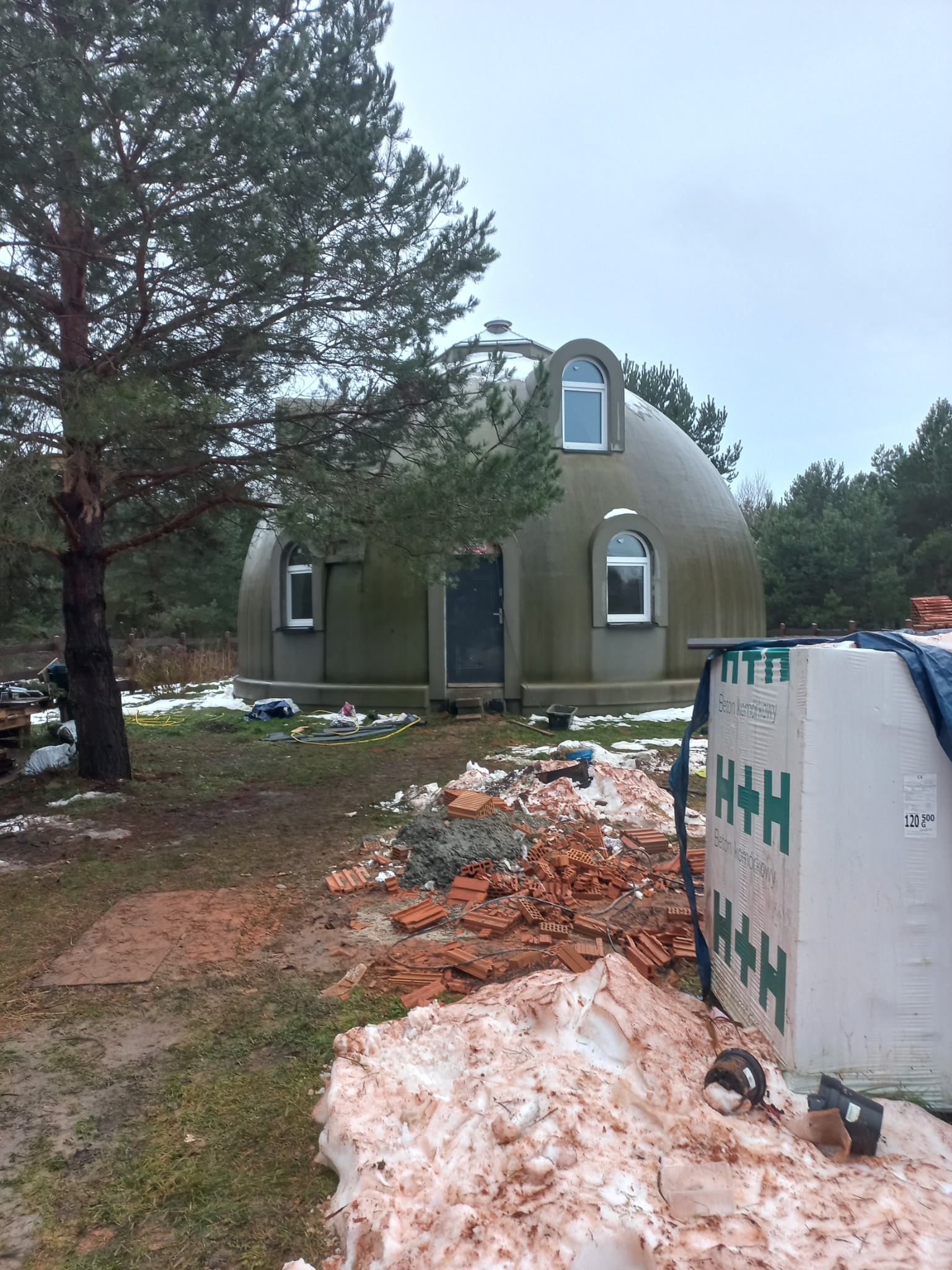 Nietypowy, okrągły dom w trakcie budowy, widoczne okna, drzwi i materiały budowlane na zewnątrz, obok drzewo iglaste i resztki śniegu.
