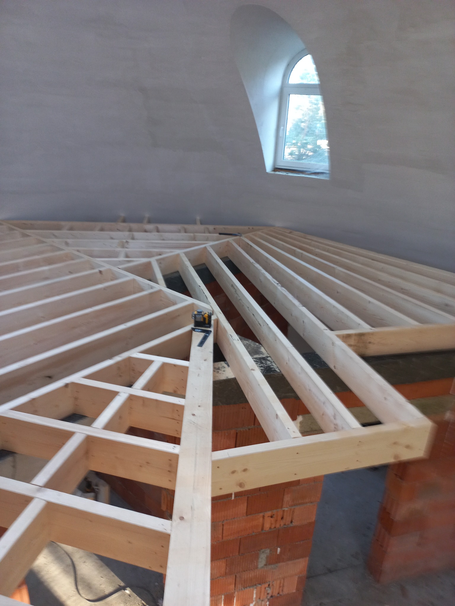 Konstrukcja drewniana dachu od wewnątrz, z widocznym oknem w szczycie i poziomicą na belce, ceglana podmurówka, surowy stan wykończenia.