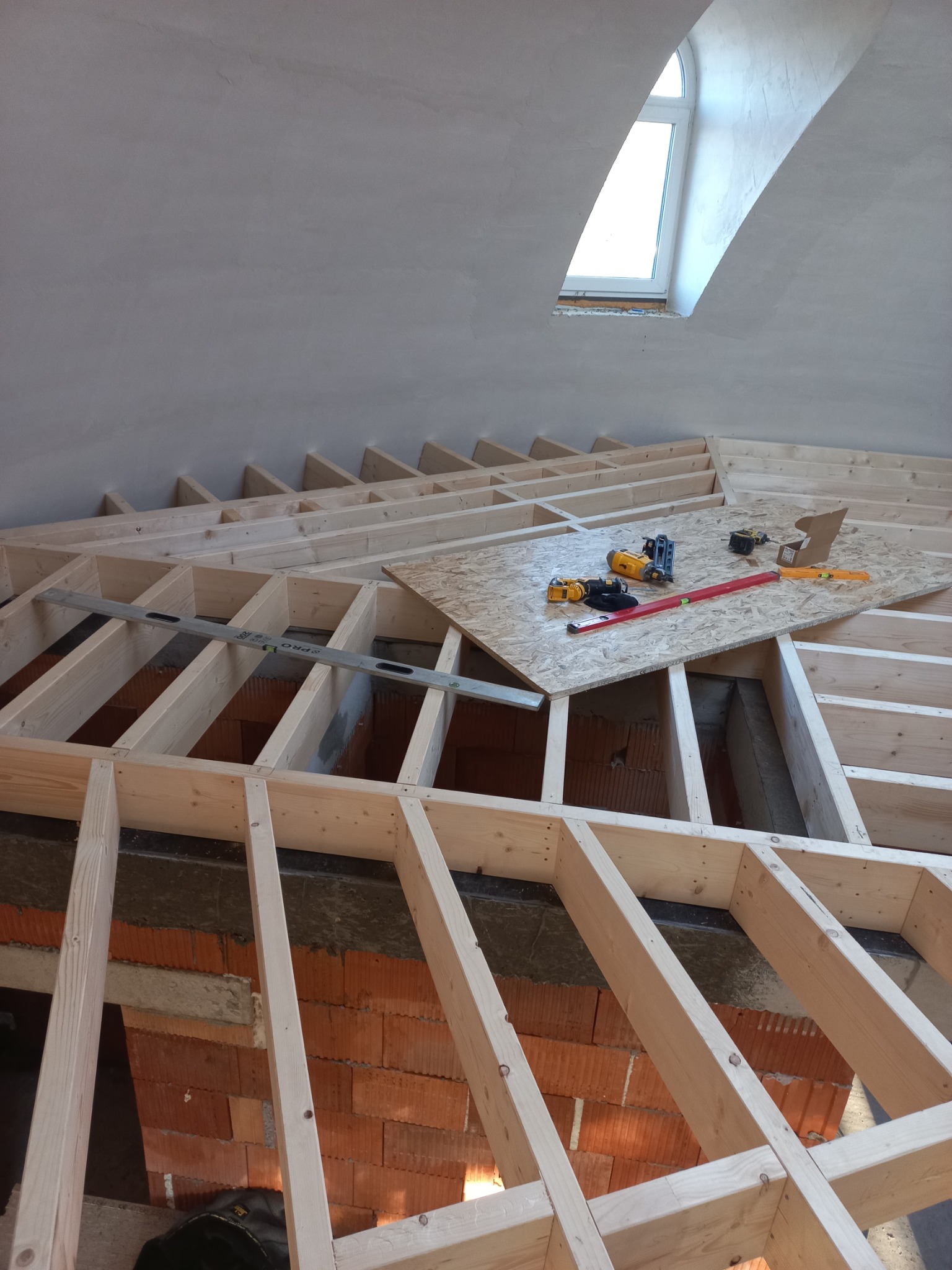 Konstrukcja drewniana poddasza z oknem połaciowym w tle. Na płycie OSB narzędzia: poziomica, wkrętarki. Widoczne cegły i surowy tynk.