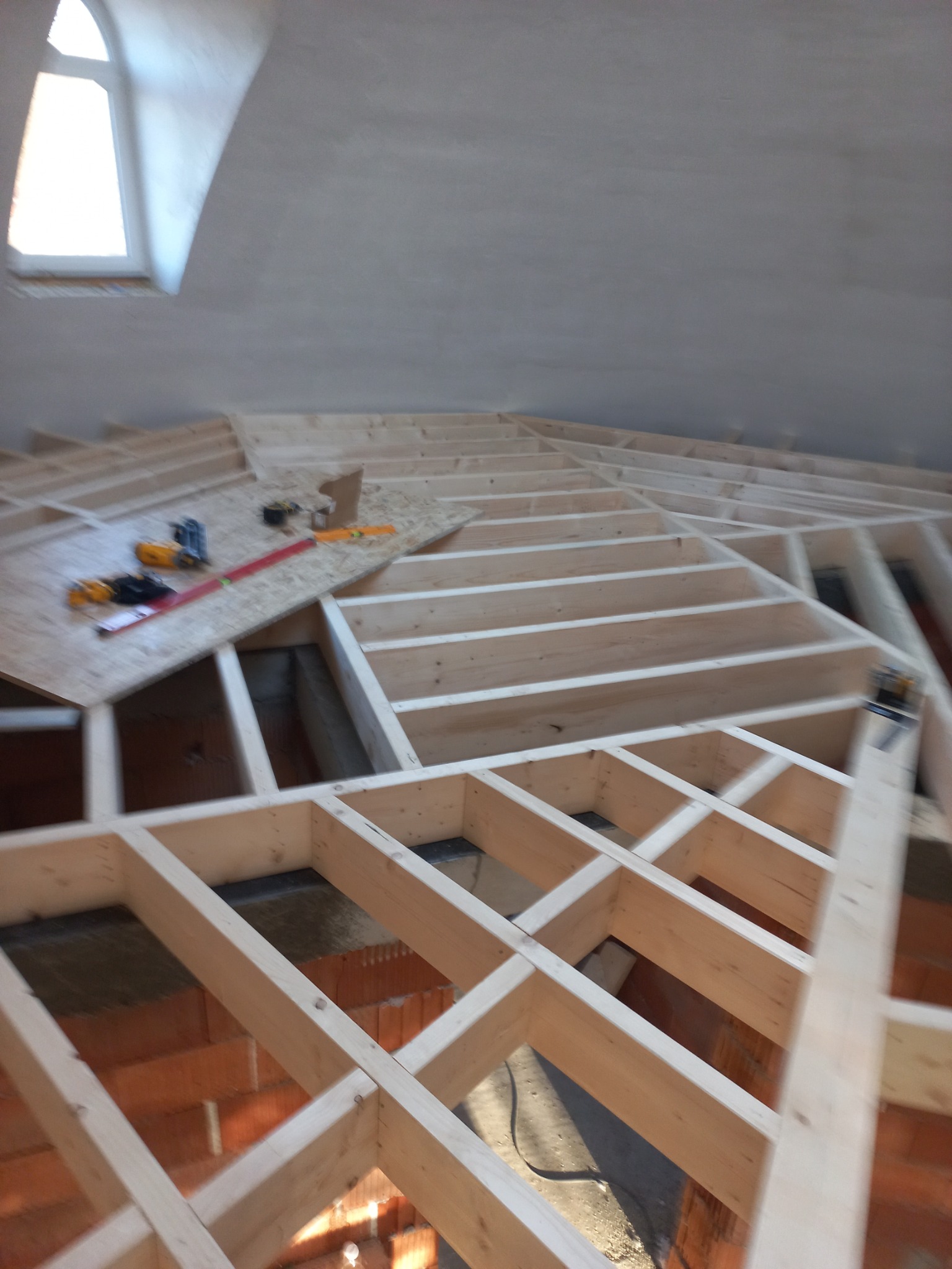 Drewniana konstrukcja stropu w trakcie budowy, widoczne narzędzia i płyta OSB, okno w tle. Elementy konstrukcji są jasne, a ceglana podstawa dodaje kontrastu.