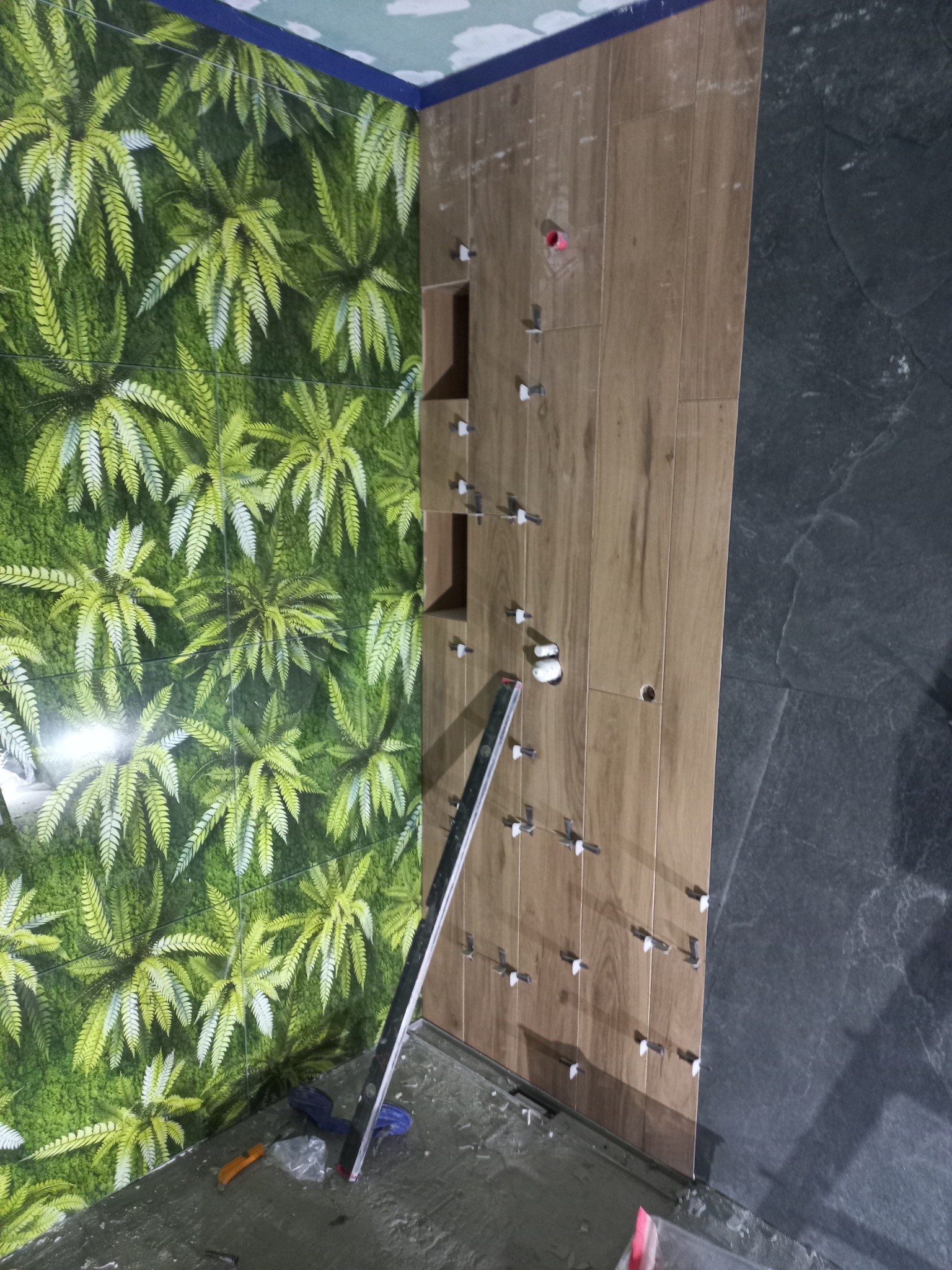 Ściana łazienki w trakcie wykańczania: część z zielonym wzorem roślinnym, część z drewnopodobnymi płytkami, poziomica i akcesoria glazurnicze.