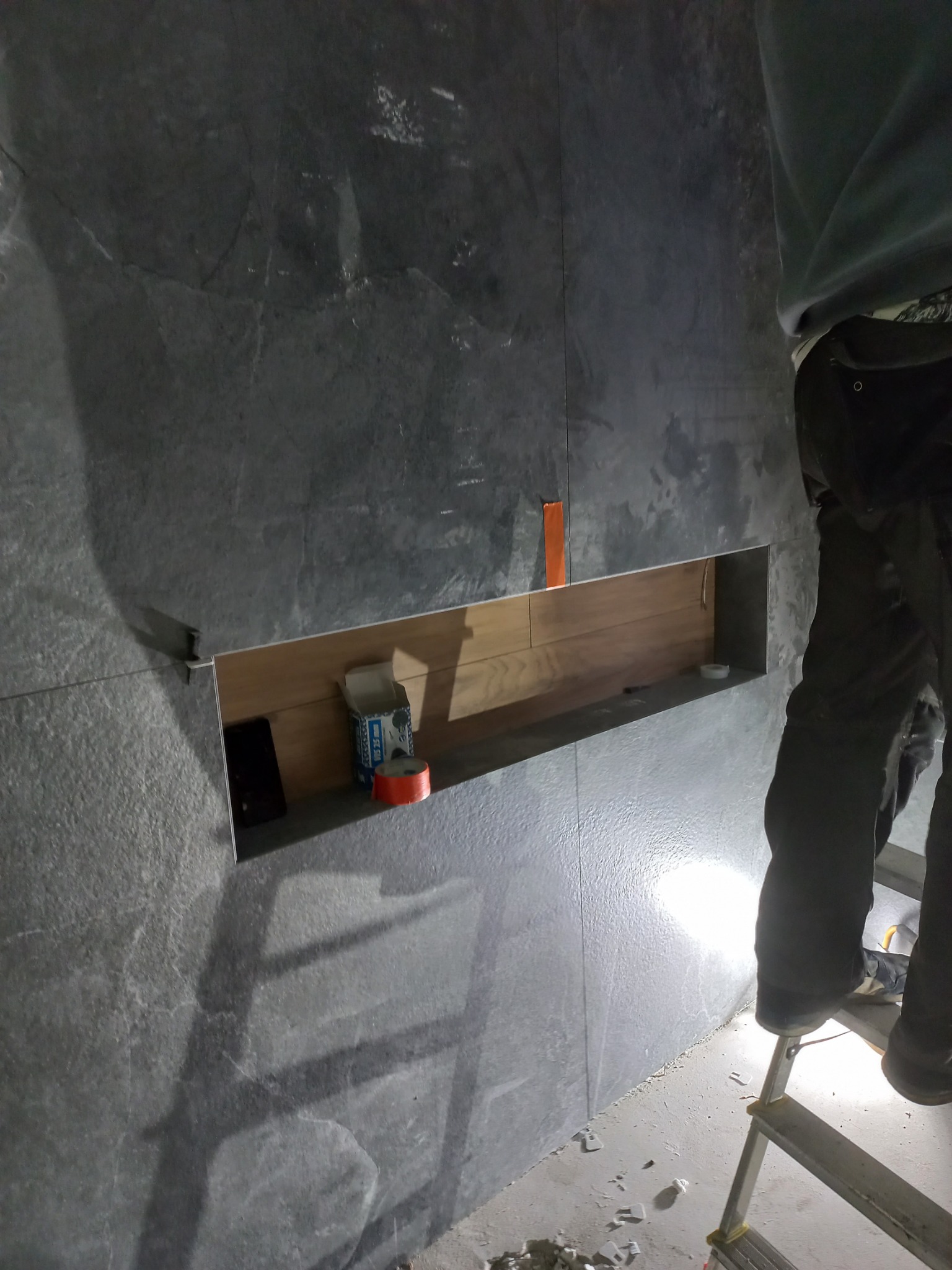 Montaż płytek imitujących kamień z wnęką wykończoną drewnem. Osoba na drabinie w trakcie pracy, widoczne narzędzia i resztki materiałów na podłodze.