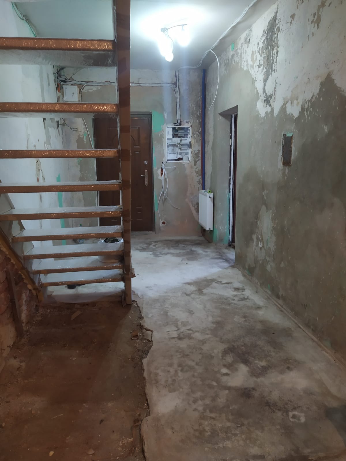 Wnętrze w trakcie remontu: surowe ściany z widocznymi śladami po farbie, drewniane schody, instalacja elektryczna i grzejnik. Widoczny stan przed wykończeniem.