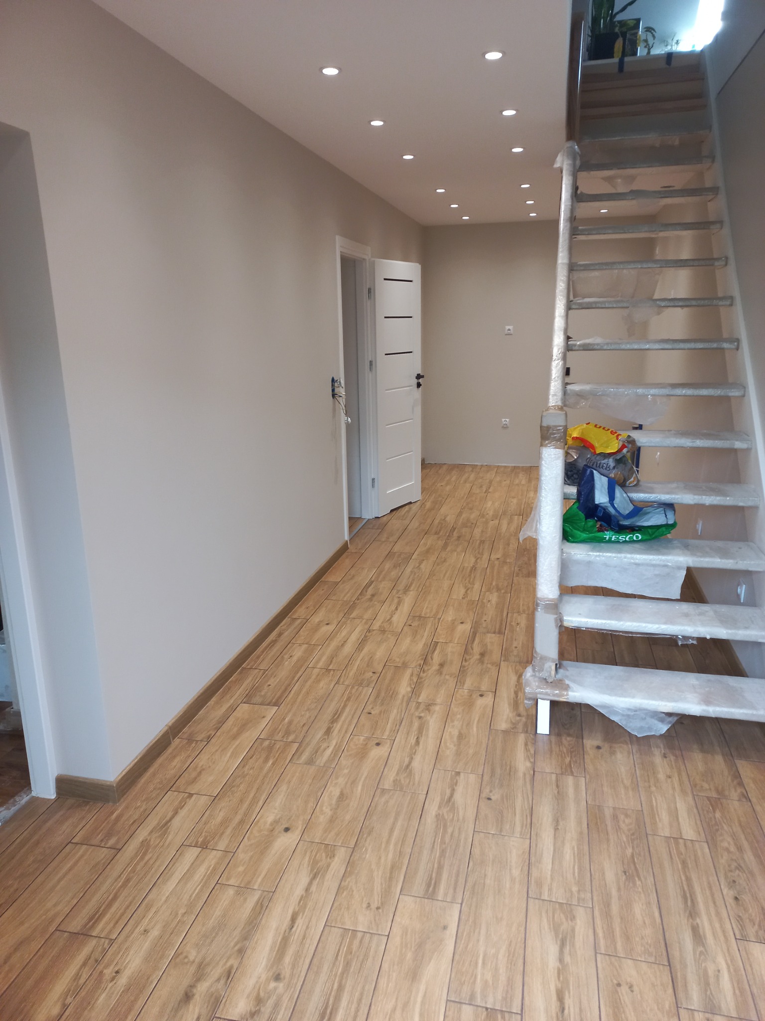 Jasne, nowo wykończone wnętrze z drewnopodobną podłogą, białymi ścianami i schodami zabezpieczonymi folią. Widoczne oświetlenie sufitowe i otwarte drzwi.