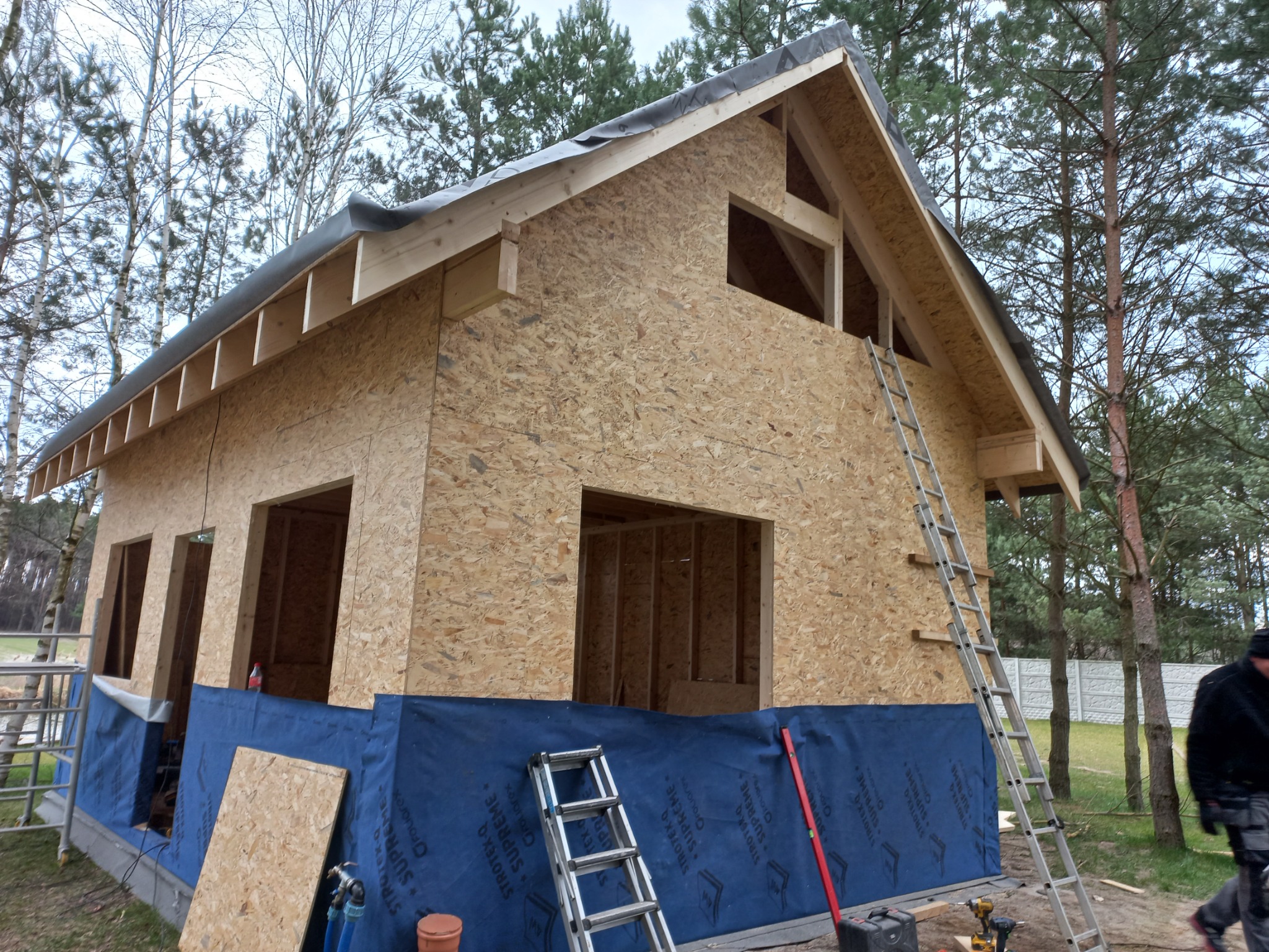 Budowa domu w trakcie realizacji: szkielet z płyt OSB, okna bez szyb, drabiny, narzędzia budowlane. Widoczne drzewa w tle.