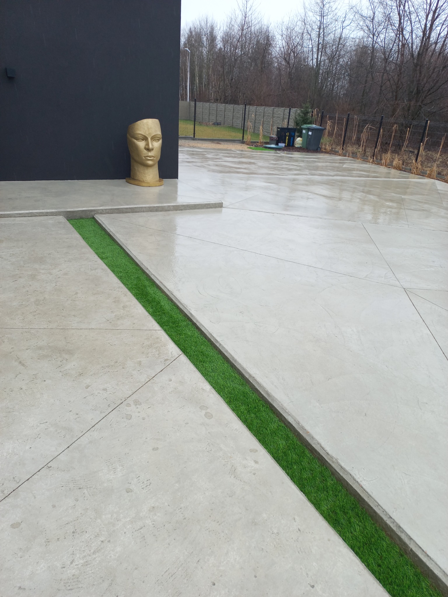 Nowoczesny betonowy taras z zielonymi wstawkami i rzeźbą w Kłobucku. Minimalistyczny design, gładka powierzchnia, widoczne geometryczne podziały i sztuczna trawa.
