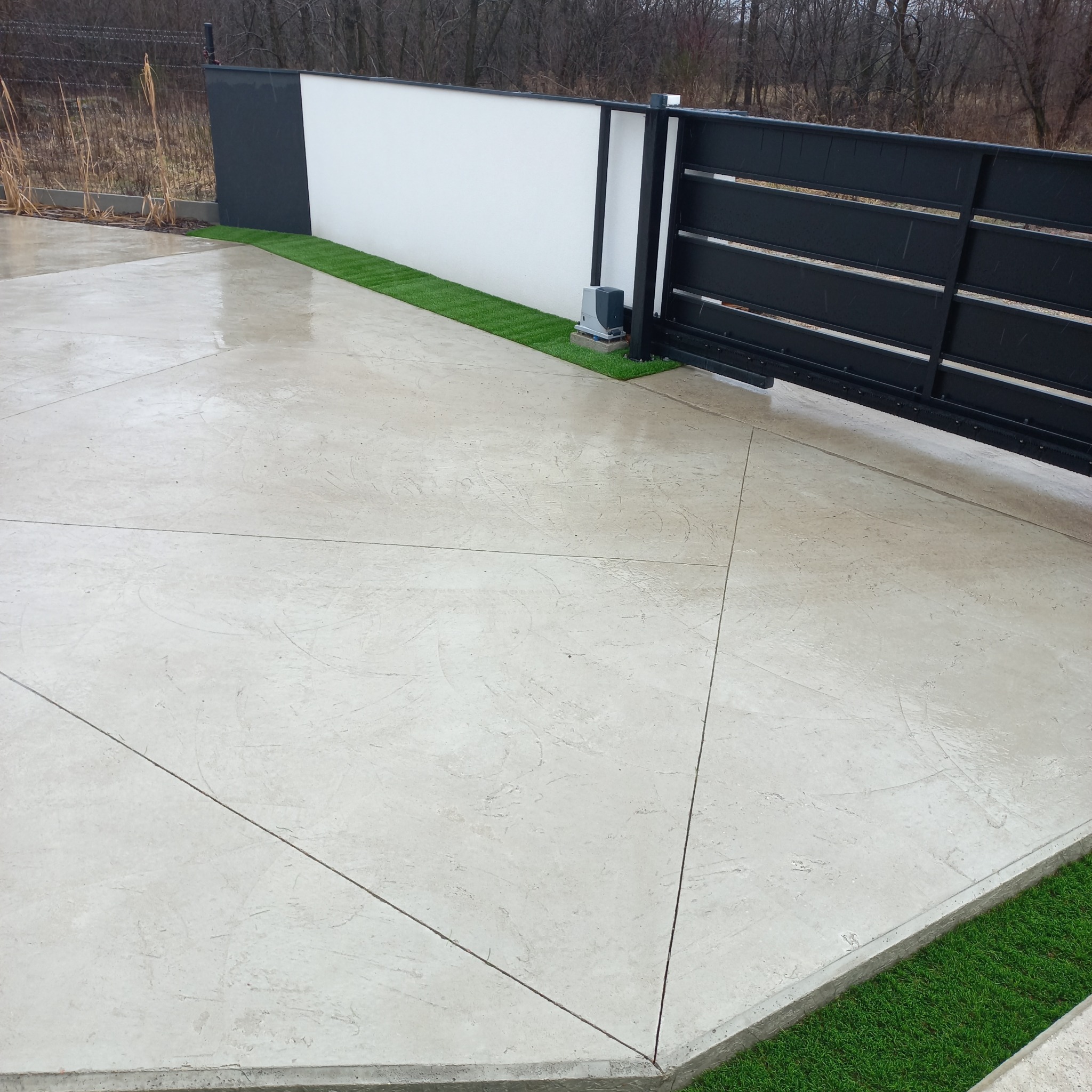 Betonowy podjazd z geometrycznym wzorem i zielonym pasem sztucznej trawy przy ogrodzeniu i bramie wjazdowej. Widoczne ślady deszczu na powierzchni.