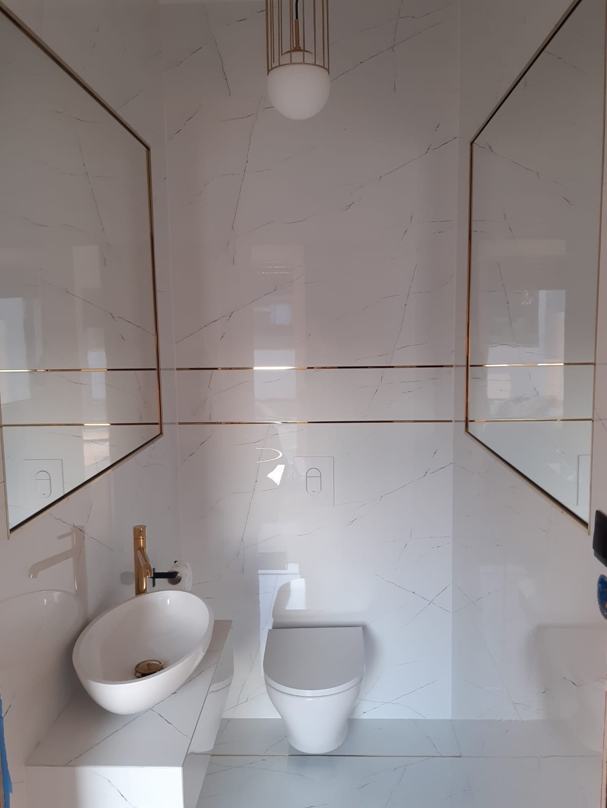 Biała łazienka z marmurowymi płytkami i złotymi akcentami: umywalka na blacie, toaleta, lustra. Minimalistyczny design, oświetlenie sufitowe.