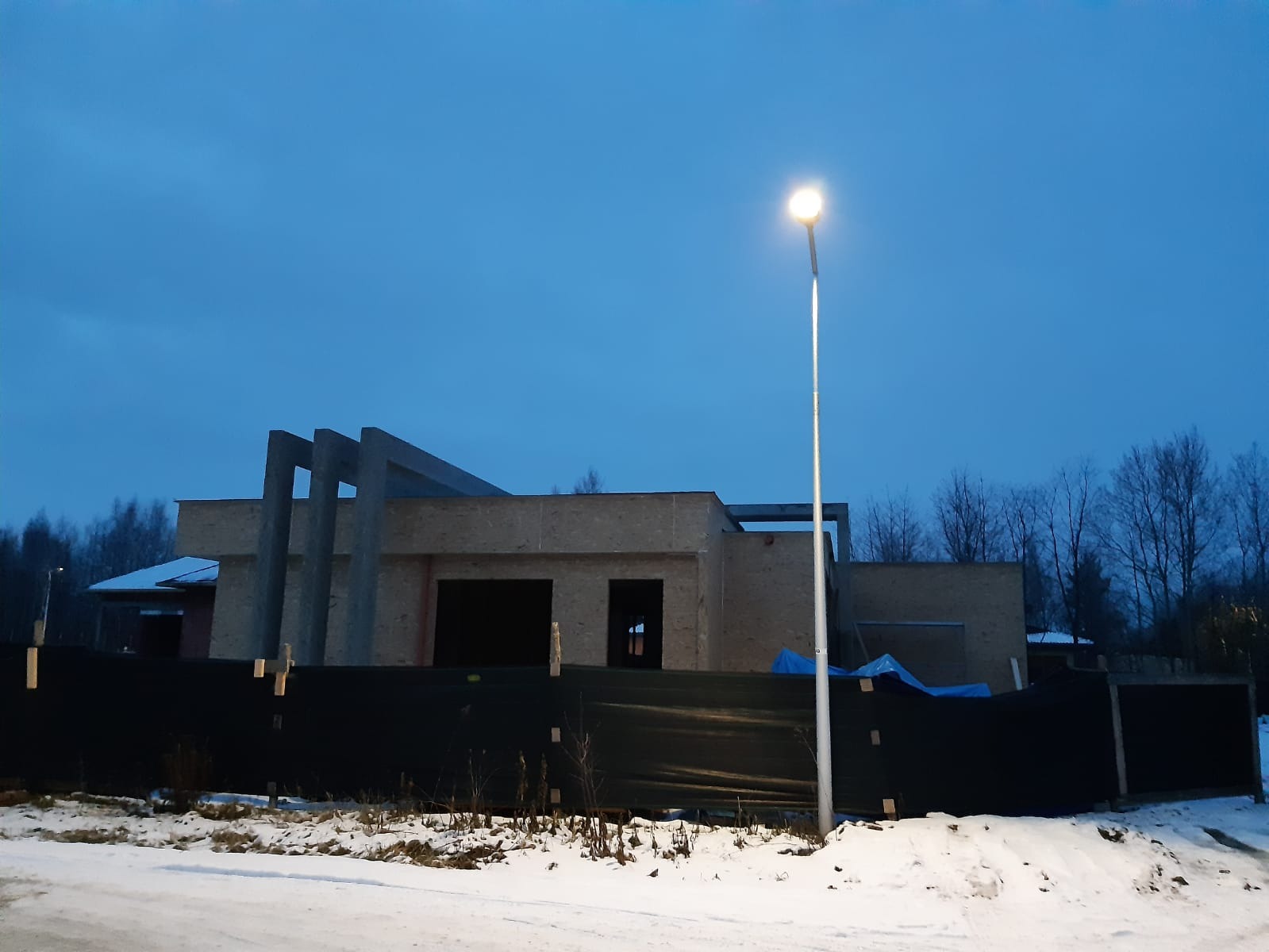 Niedokończony dom z betonu komórkowego, otoczony czarnym płotem i śniegiem, oświetlony przez latarnię uliczną na tle błękitnego, zimowego nieba.