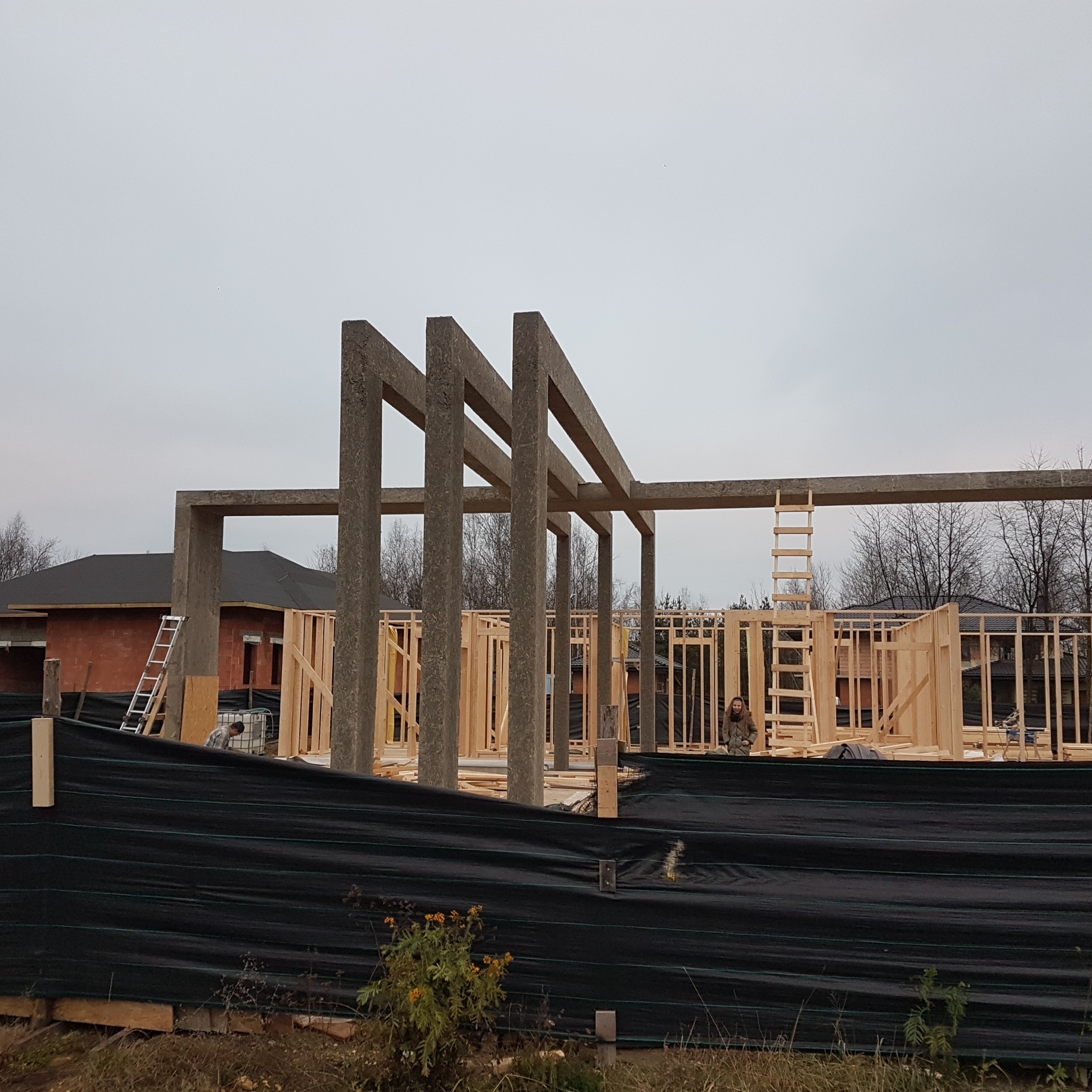 Surowa konstrukcja domu w trakcie budowy: drewniane szkielety ścian i betonowe słupy na tle pochmurnego nieba. Widoczne narzędzia i drabina.