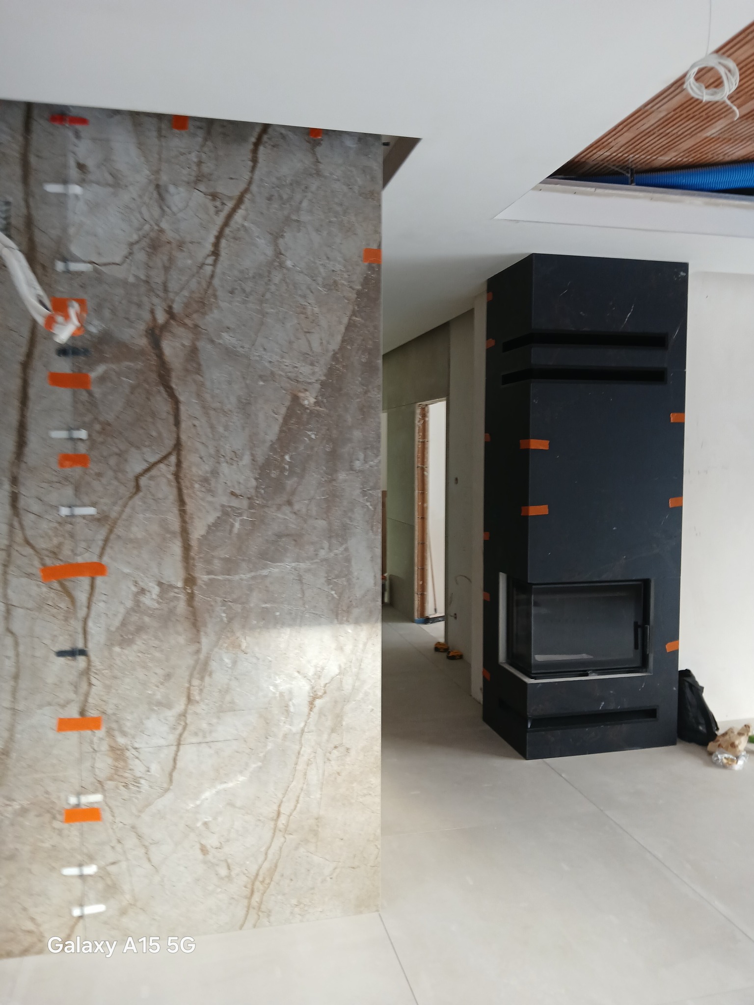 Marmurowa ściana z poziomowaniem i czarny kominek z taśmą malarską w surowym wnętrzu. Widoczne elementy instalacji pod sufitem.