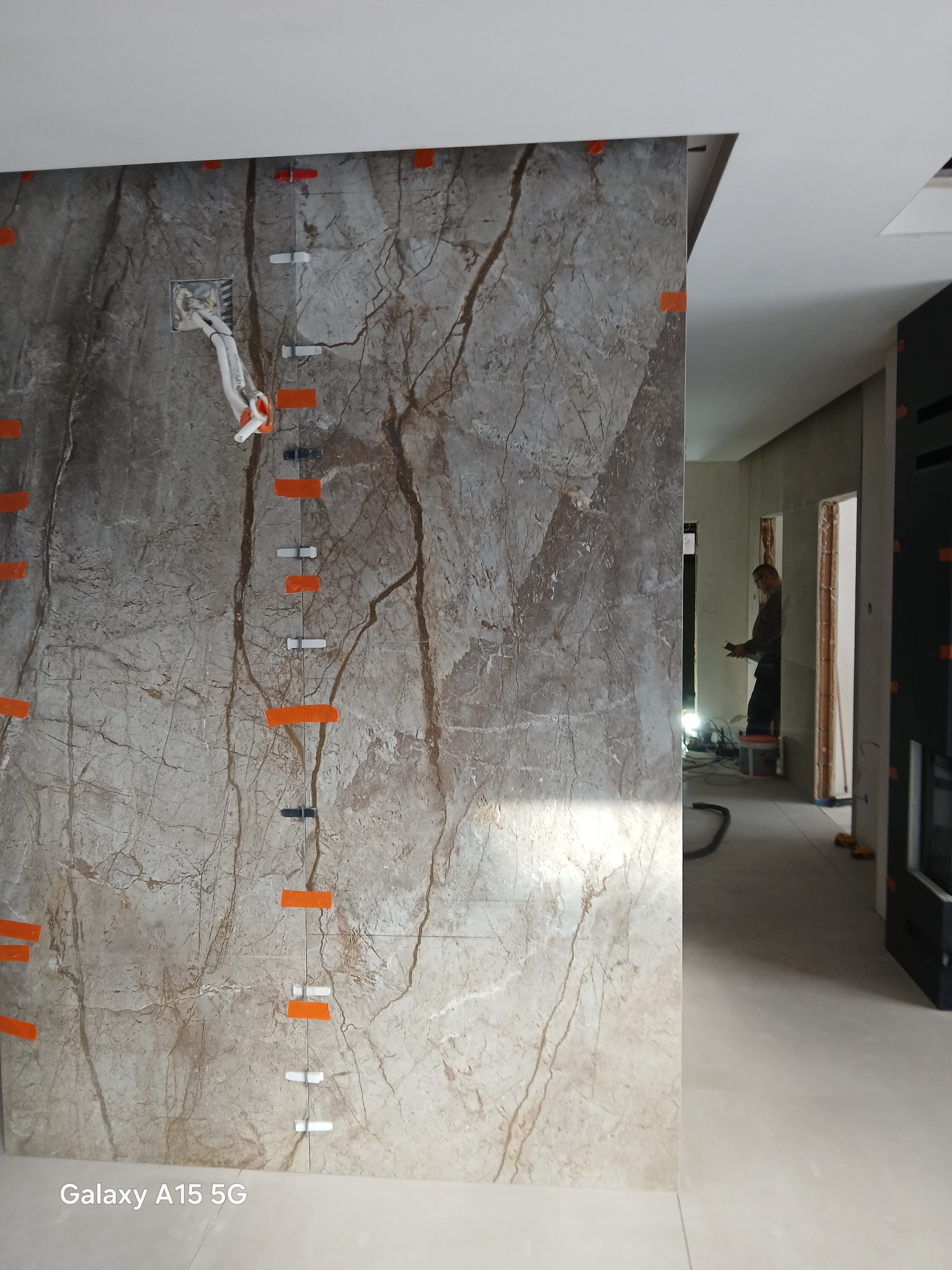 Montaż marmurowych płytek na ścianie z widocznymi elementami poziomowania i gniazdkiem elektrycznym. W tle widoczna postać pracownika.