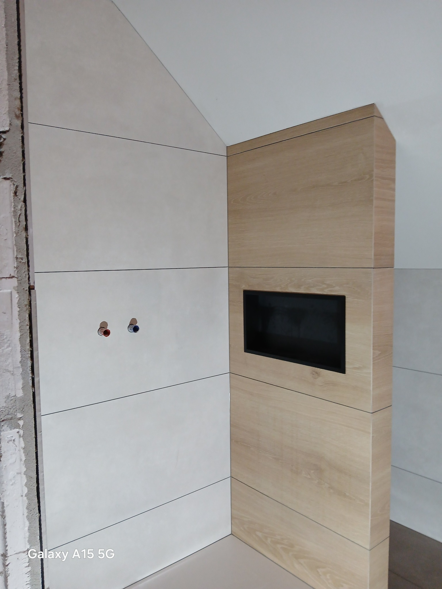 Ściana łazienki z jasnymi płytkami i drewnopodobną zabudową z wnęką na TV, widoczne przyłącza wodne. Częściowo wykończone, surowe krawędzie.
