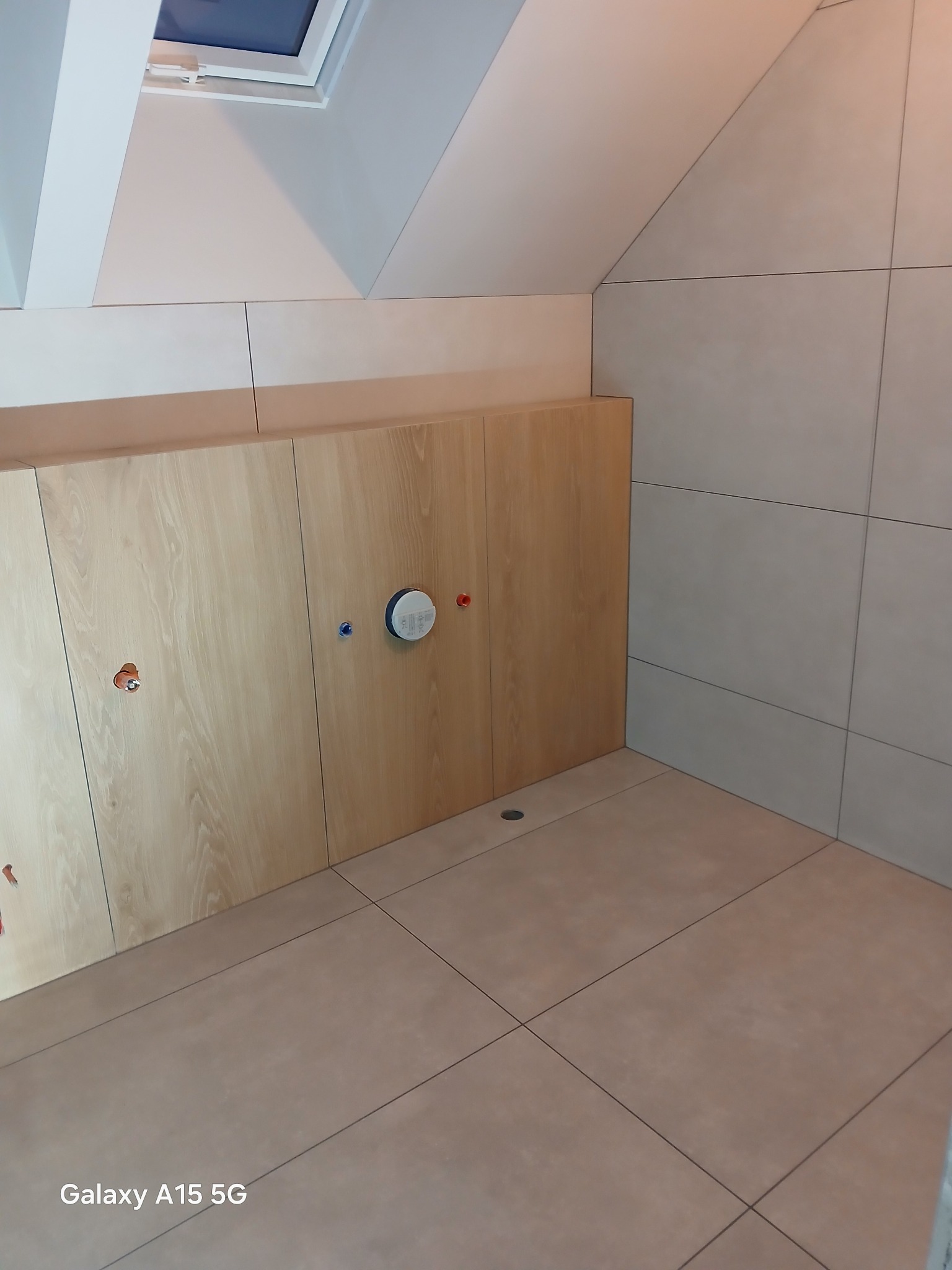 Wykończona łazienka na poddaszu: beżowe płytki na ścianach i podłodze, zabudowa z drewnianymi frontami, widoczne przyłącza wodne i okno dachowe.