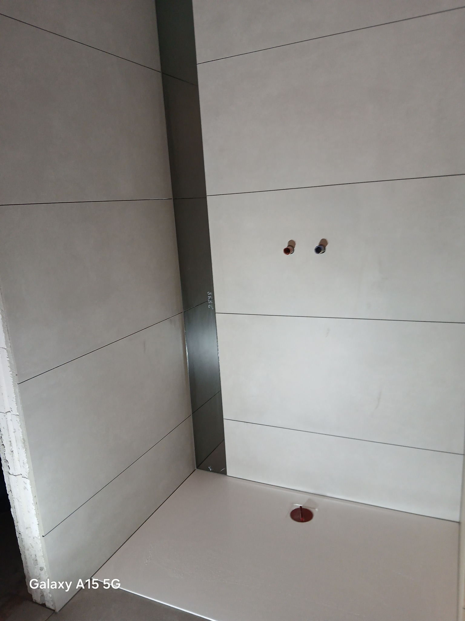 Wykończona kabina prysznicowa z jasnymi, poziomymi płytkami i ciemną listwą. Widoczne przyłącza wodne i odpływ. Minimalistyczny design.