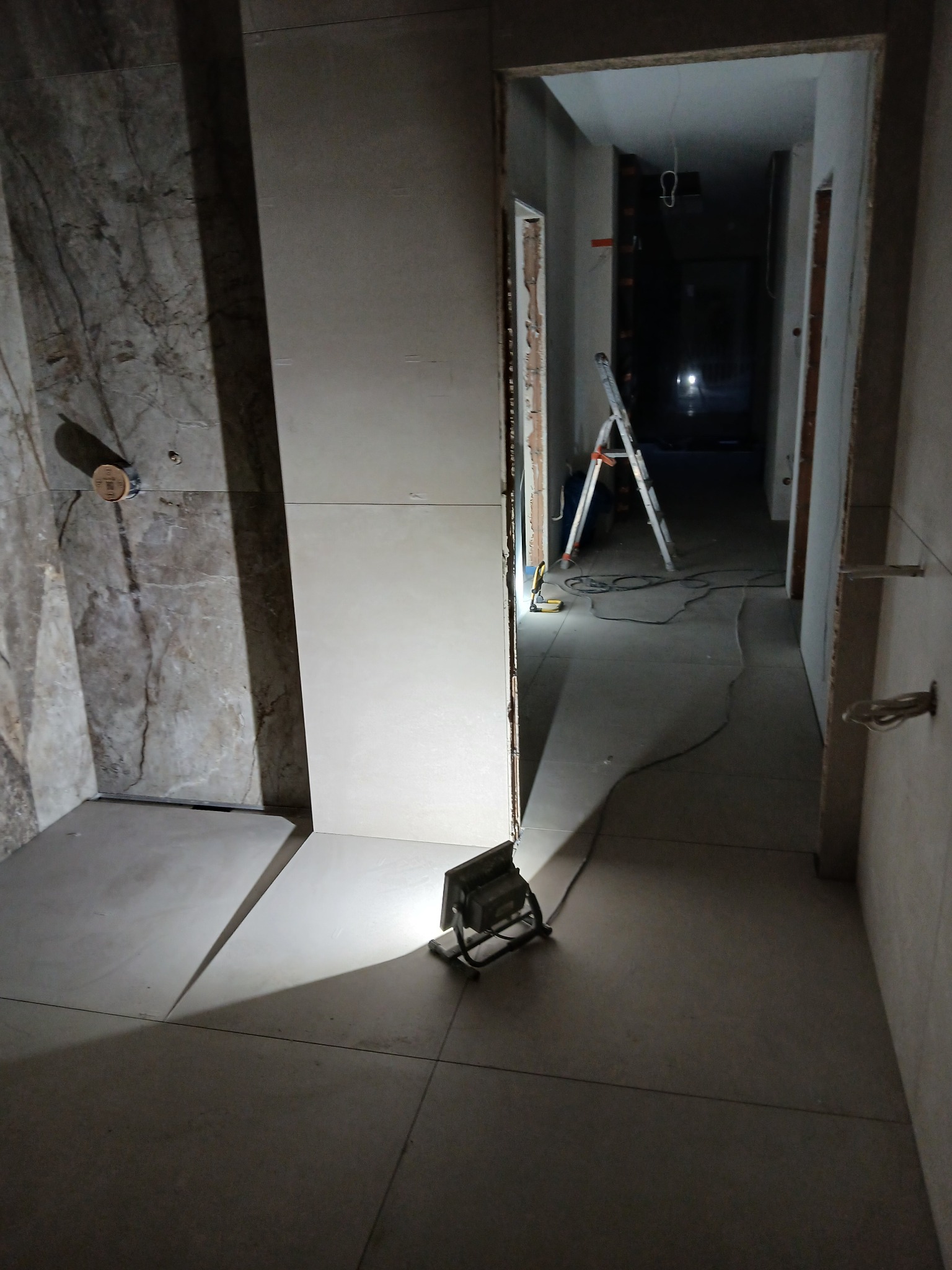 Wnętrze łazienki w trakcie wykańczania: marmurowe płytki na ścianie, jasne płytki na podłodze, lampa budowlana i drabina w tle. Widoczne niedokończone prace.