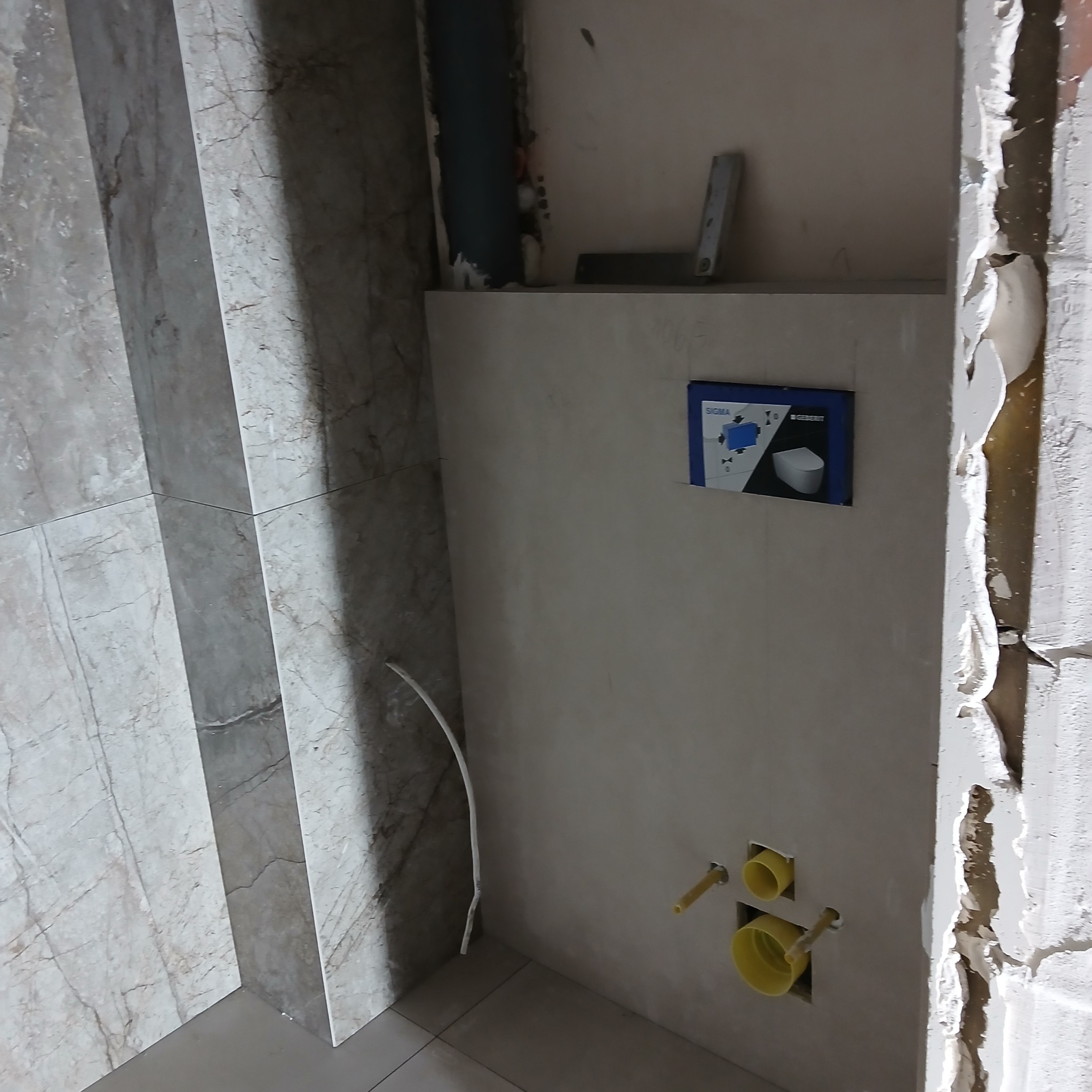 Montaż stelaża podtynkowego WC z widocznymi przyłączami i częściowo położonymi marmurowymi płytkami. Prace w toku, surowy stan wykończenia.