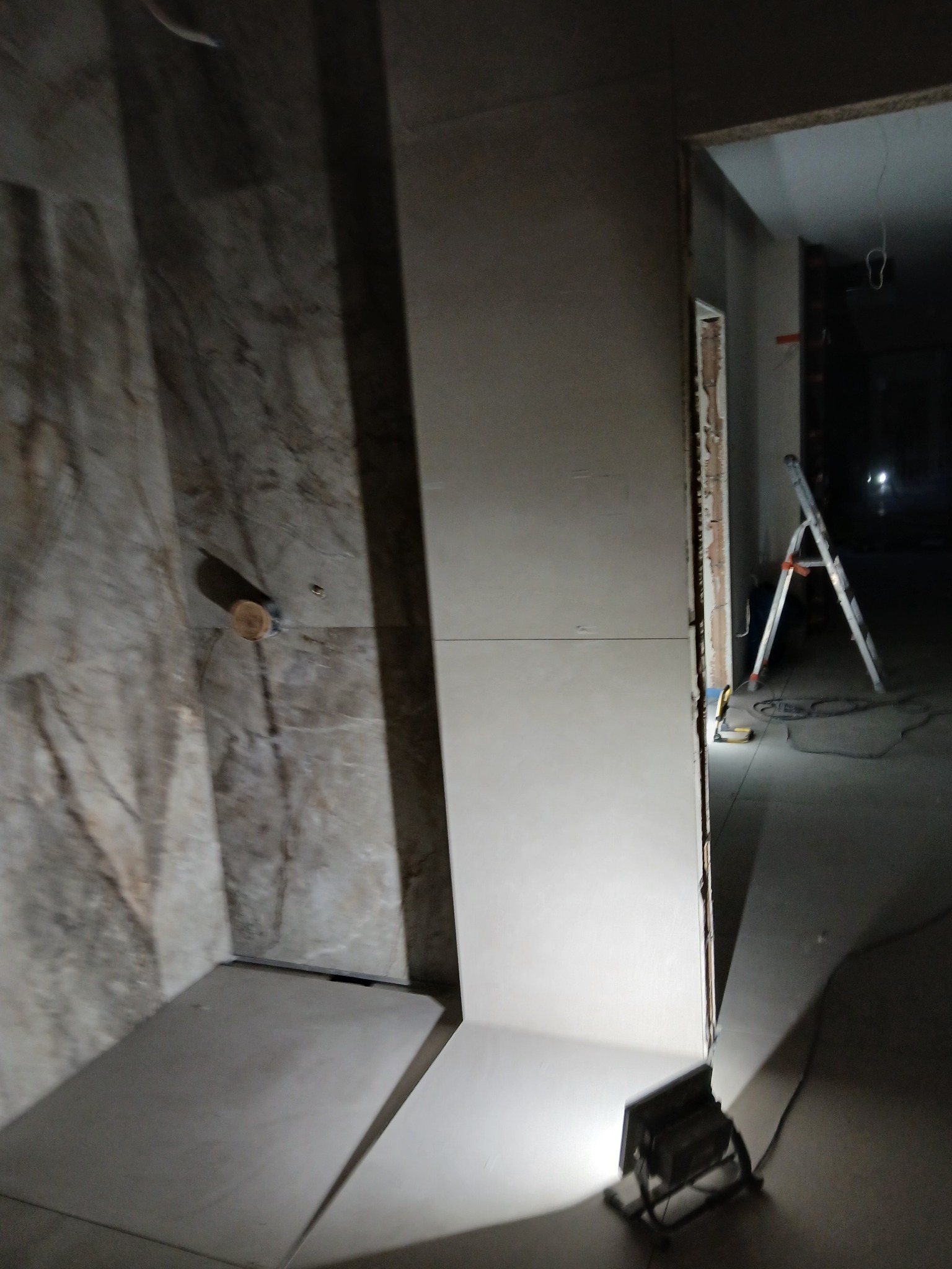 Wnętrze łazienki w trakcie prac wykończeniowych: częściowo położone płytki imitujące marmur, przygotowane ściany, drabina i lampa budowlana w tle.