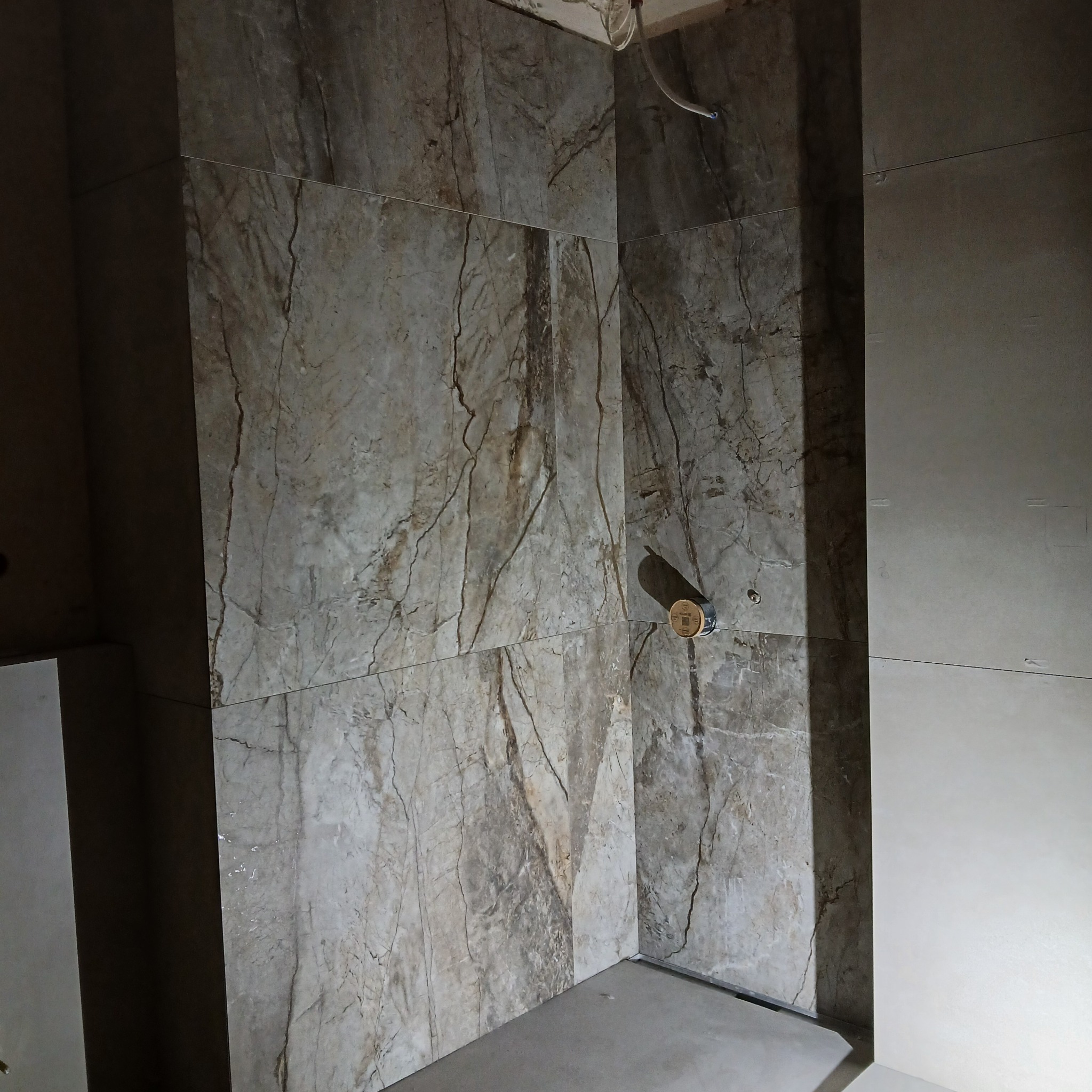 Wykończona ściana łazienki z marmurowymi płytkami, widoczny odpływ liniowy i fragment instalacji. Płyty w odcieniach szarości i beżu z wyraźnym wzorem żyłkowania.