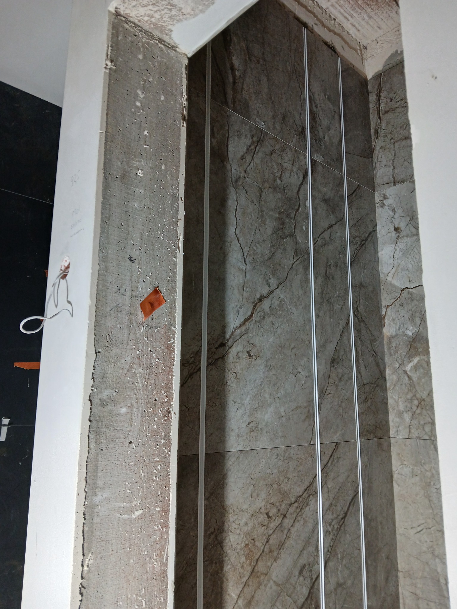 Wnęka z marmurowymi płytkami, widoczne profile aluminiowe i surowy beton. Etap prac wykończeniowych, detale montażu płytek.