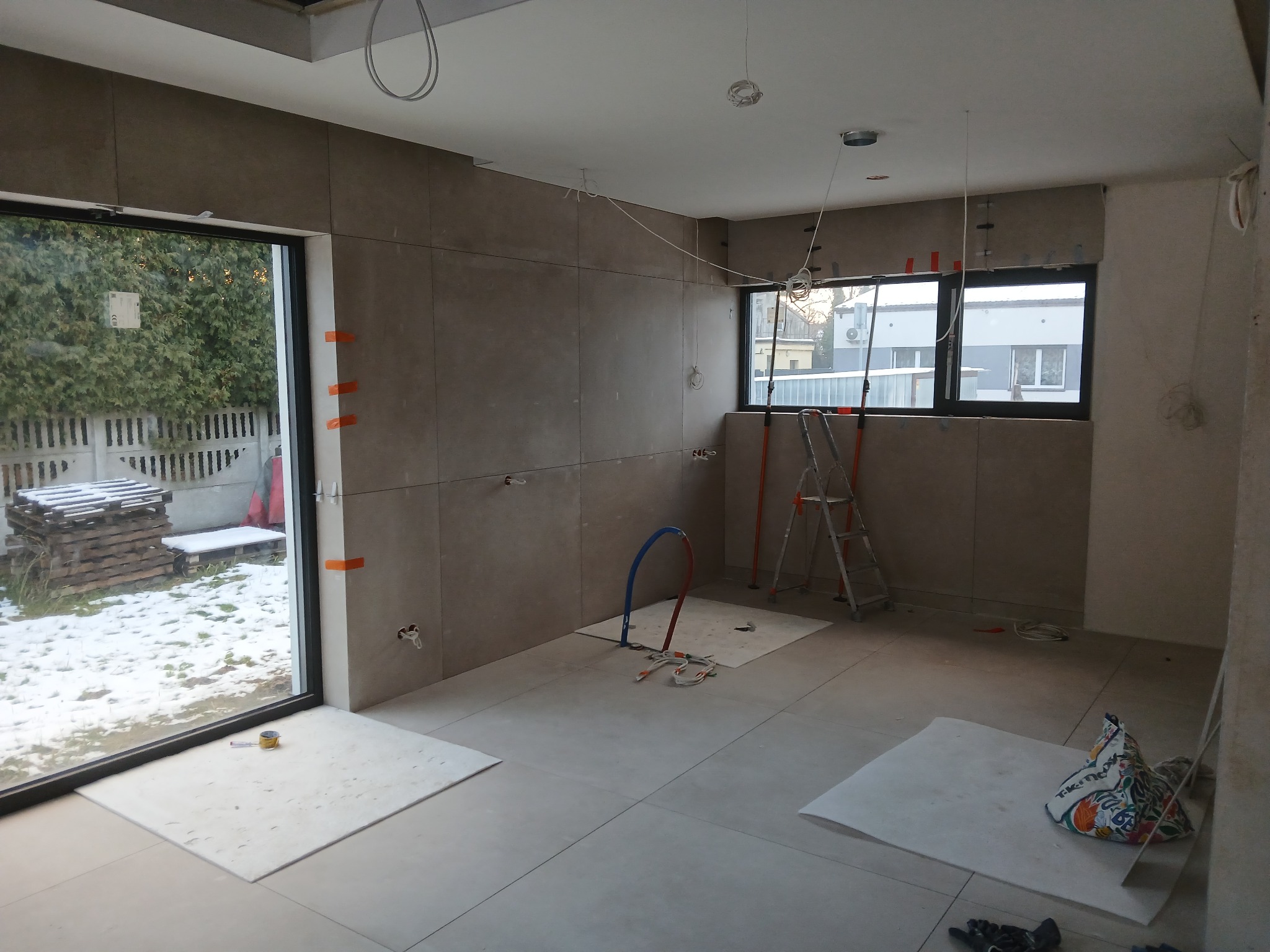 Wnętrze w trakcie prac wykończeniowych: ułożone płytki na ścianach i podłodze, widoczne instalacje, drabina i okno z widokiem na zimowy ogród.