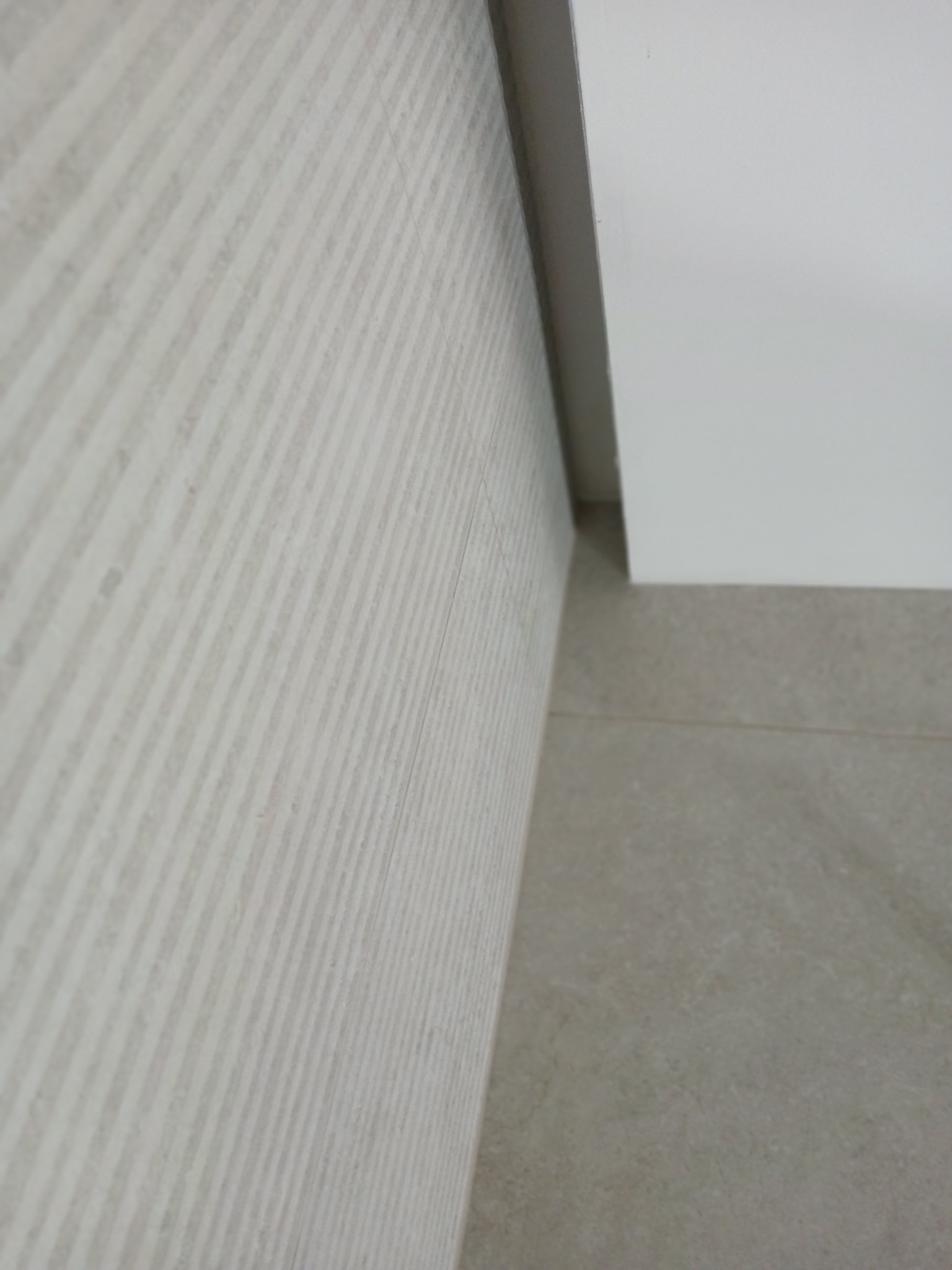 Ściana wyłożona jasnymi, prążkowanymi płytkami ceramicznymi, z widocznym fragmentem sufitu i podłogi z płytek w odcieniach beżu. Minimalistyczne wykończenie.