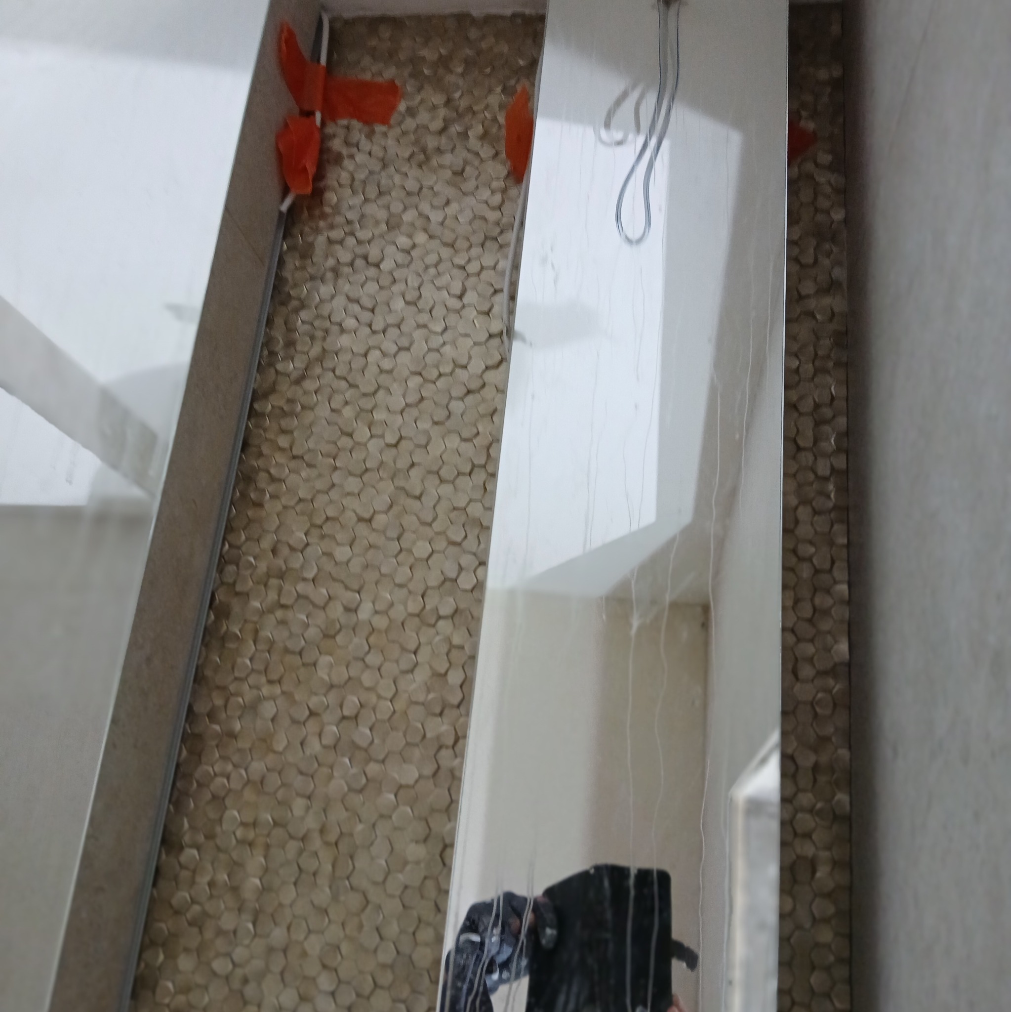 Układanie mozaiki heksagonalnej w łazience, widoczne płytki przytrzymywane pomarańczowymi klipsami, odbicie narzędzi glazurniczych w lustrzanej powierzchni.