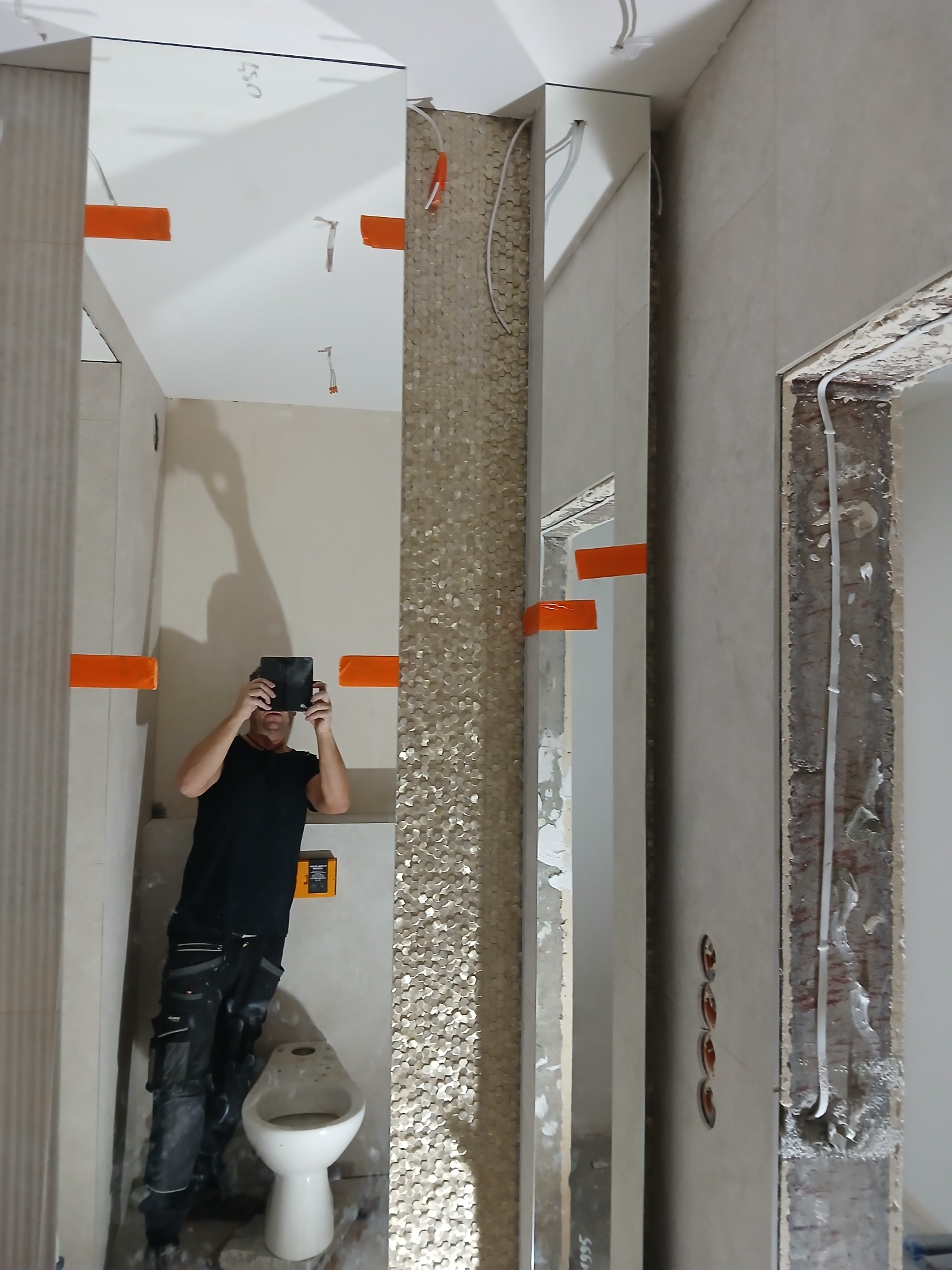 Łazienka w trakcie remontu: mozaika na ścianie, lustra z pomarańczową taśmą, instalacja elektryczna. Osoba robi zdjęcie postępów prac.