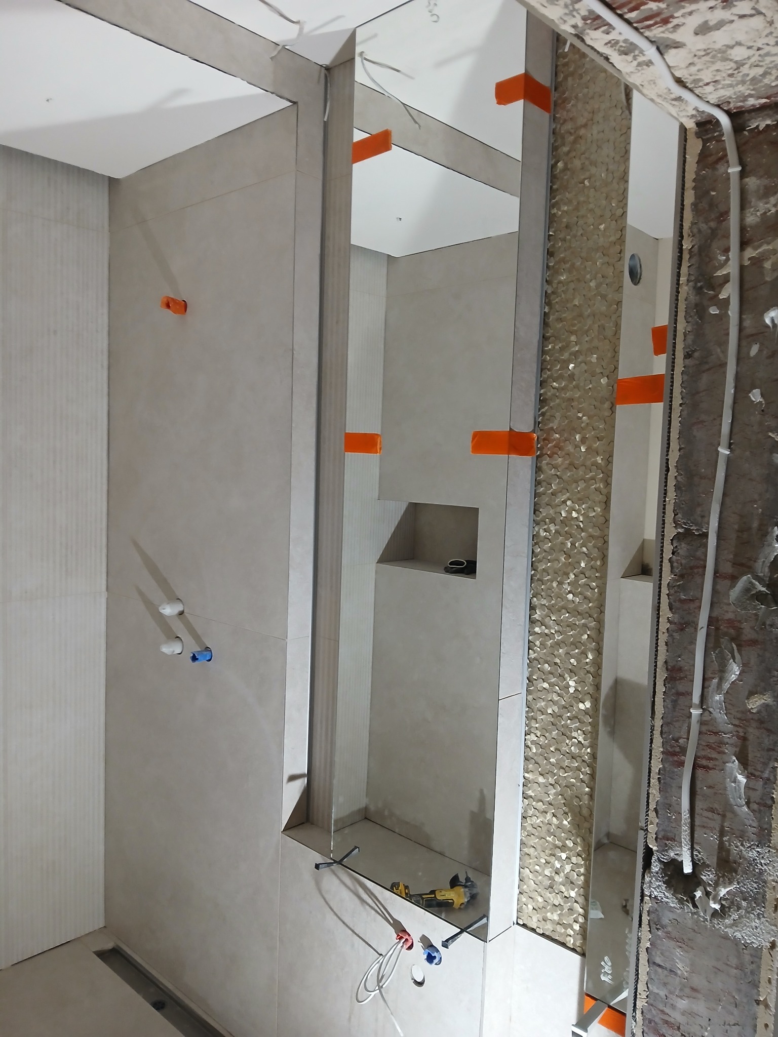 Wnętrze łazienki w trakcie wykańczania: beżowe płytki, mozaika na ścianie, widoczne narzędzia i instalacje. Pomarańczowe kliny do płytek.