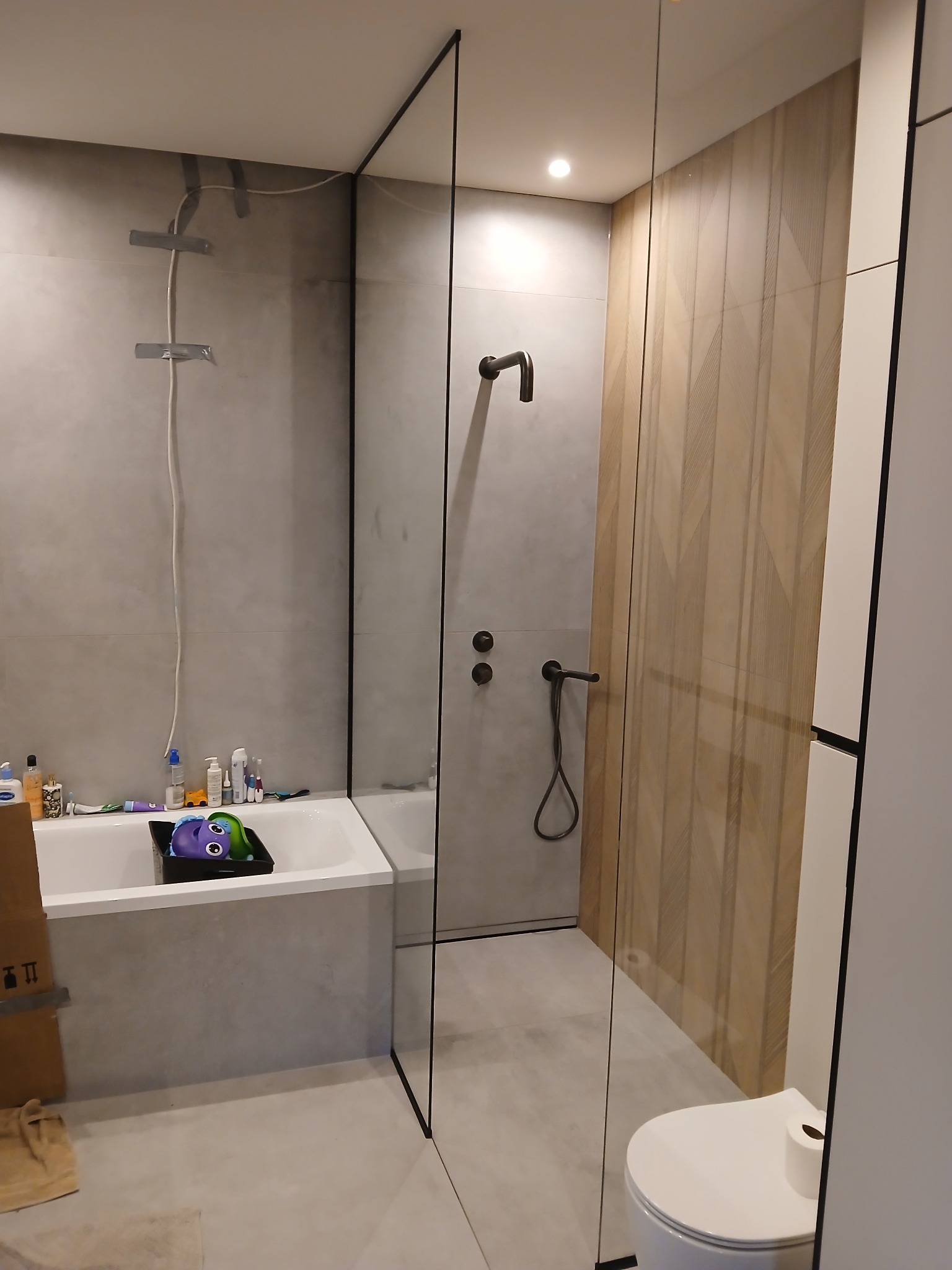 Nowoczesna łazienka z kabiną typu walk-in, wykończona płytkami w odcieniach szarości i drewna. Widoczna wanna z akcesoriami i toaleta. Minimalistyczny design.