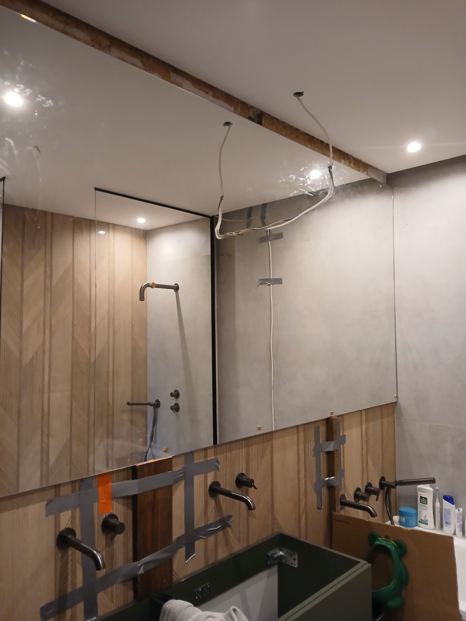 Łazienka w trakcie montażu armatury: widoczne przytwierdzone taśmą baterie, lustro z przewodami, płytki imitujące drewno i fragment umywalki. Etap prac wykończeniowych.