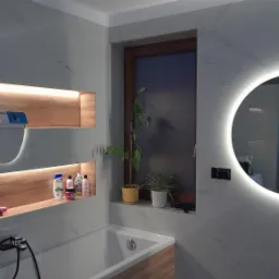 Wykończona łazienka z marmurowymi płytkami, podświetlanymi niszami z drewnianym wykończeniem, okrągłym lustrem z podświetleniem LED i roślinami w doniczkach przy oknie.