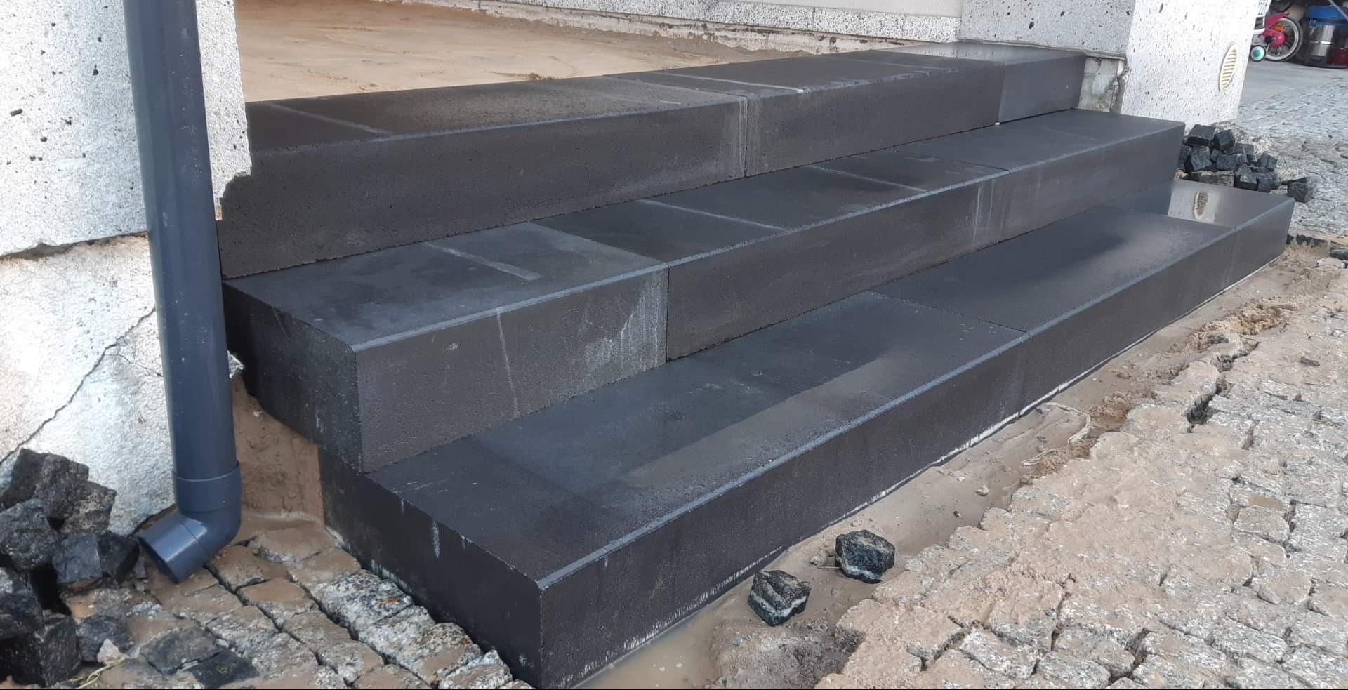 Nowe, ciemne, betonowe schody wejściowe z kostki brukowej w trakcie montażu, widoczny fragment rynny spustowej i ułożona kostka brukowa wokół.