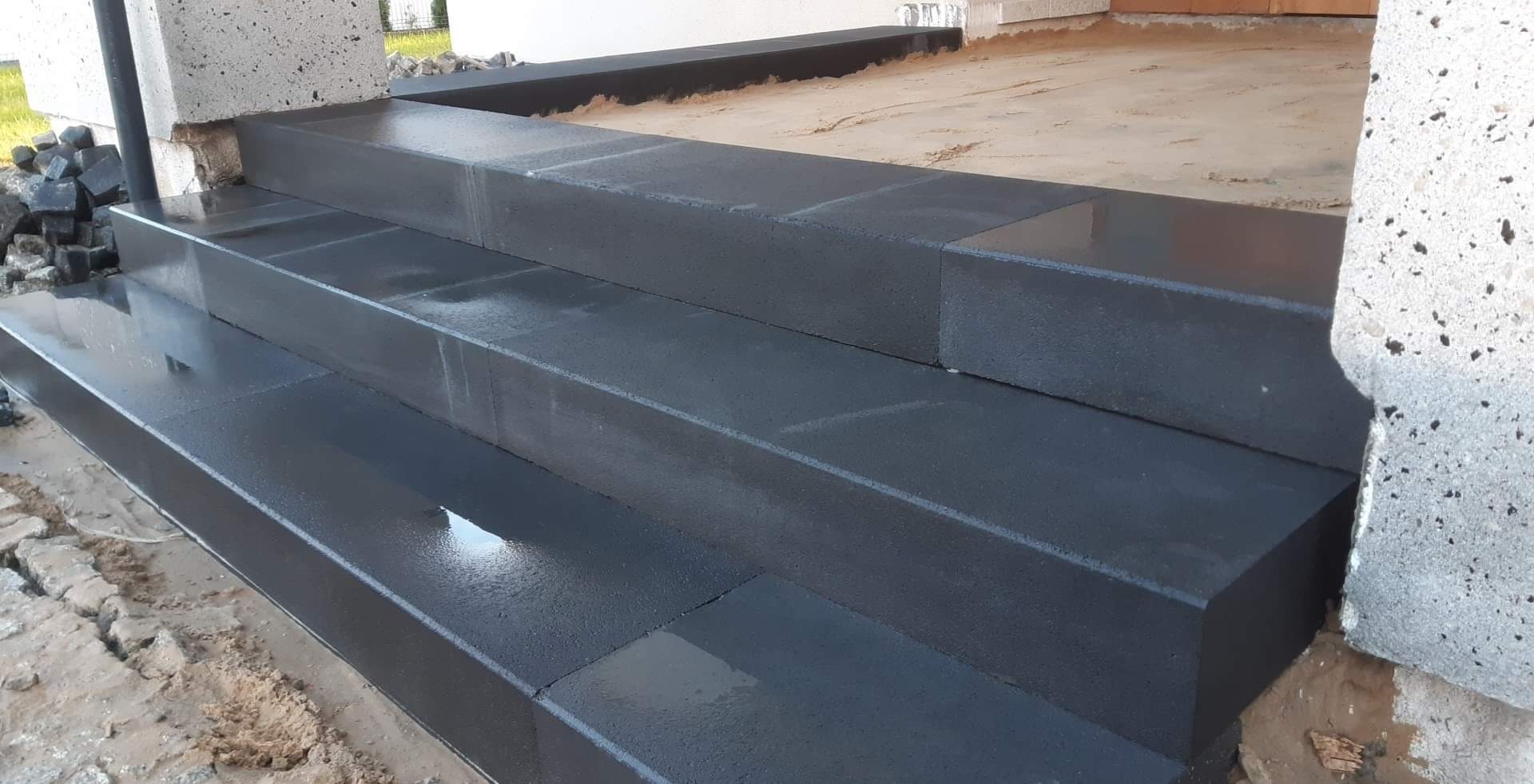 Nowe, ciemnoszare, betonowe schody wejściowe, świeżo po ułożeniu, z widocznymi drobnymi kamieniami kostki brukowej obok i śladami wilgoci.