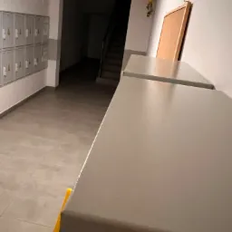 Szare szafki w korytarzu bloku mieszkalnego, żółty znak ostrzegawczy o śliskiej podłodze, skrzynki pocztowe w tle.