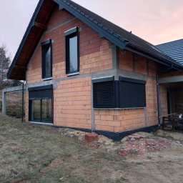 MATEUSZ KALISZUK TechRol - Surowy dom z cegły z zamontowanymi czarnymi osłonami okiennymi. Widoczne niedokończone otoczenie i elementy budowlane. Ujęcie z dołu, pochmurne niebo.