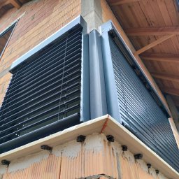 MATEUSZ KALISZUK TechRol - Szare żaluzje fasadowe zamontowane na rogu budynku z cegły, widoczne elementy konstrukcyjne dachu. Ujęcie z dołu, podkreślające nowoczesny design.