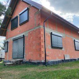 MATEUSZ KALISZUK TechRol - Dom w trakcie budowy z zamontowanymi, czarnymi roletami zewnętrznymi na oknach. Widoczne palety i elementy instalacji. Ujęcie z dołu, w tle błękitne niebo.