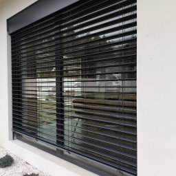 MATEUSZ KALISZUK TechRol - Eleganckie, grafitowe żaluzje zewnętrzne zamontowane w oknie nowoczesnego domu. Widoczne białe kamienie dekoracyjne i krzewy u podstawy ściany.