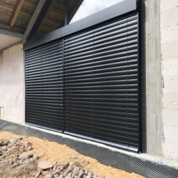 MATEUSZ KALISZUK TechRol - Ciemne, poziome lamele na elewacji budynku w trakcie budowy. Widoczne bloczki betonowe i fragment dachu. Ujęcie z dołu, podkreślające nowoczesny wygląd.