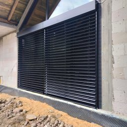 MATEUSZ KALISZUK TechRol - Ciemne, poziome lamele okienne zamontowane w surowym budynku z betonu komórkowego. Widoczne zabezpieczenie fundamentów i piasek.