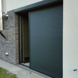 MATEUSZ KALISZUK TechRol - Zamknięta, grafitowa osłona okienna na parterze domu z szarej cegły i białego tynku. Widoczny fragment trawnika i skrzynka narzędziowa.